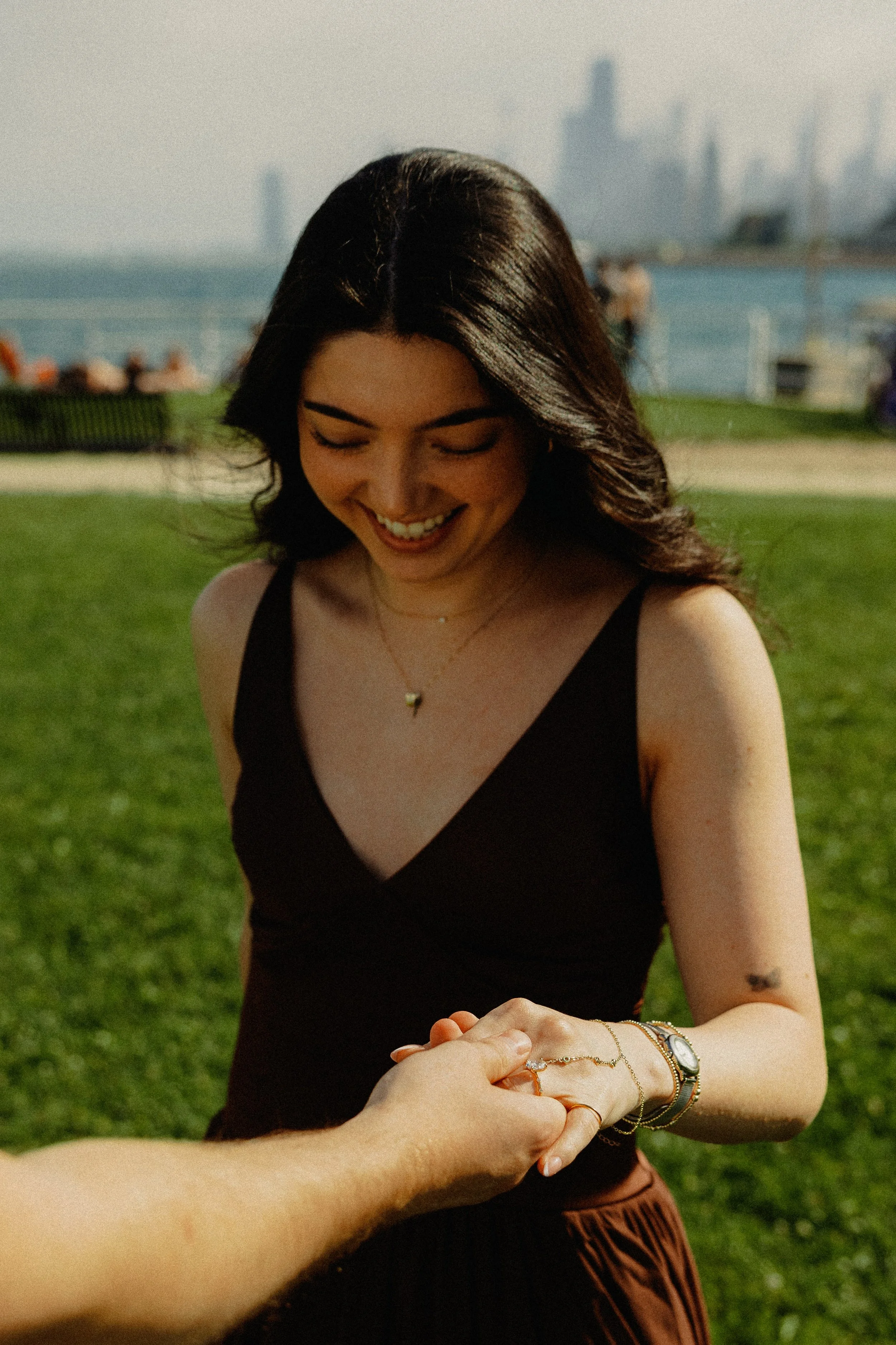 A young woman with long dark hair smiling and looking down outdoors, holding someone's hand, with a city skyline and waterfront in the background.