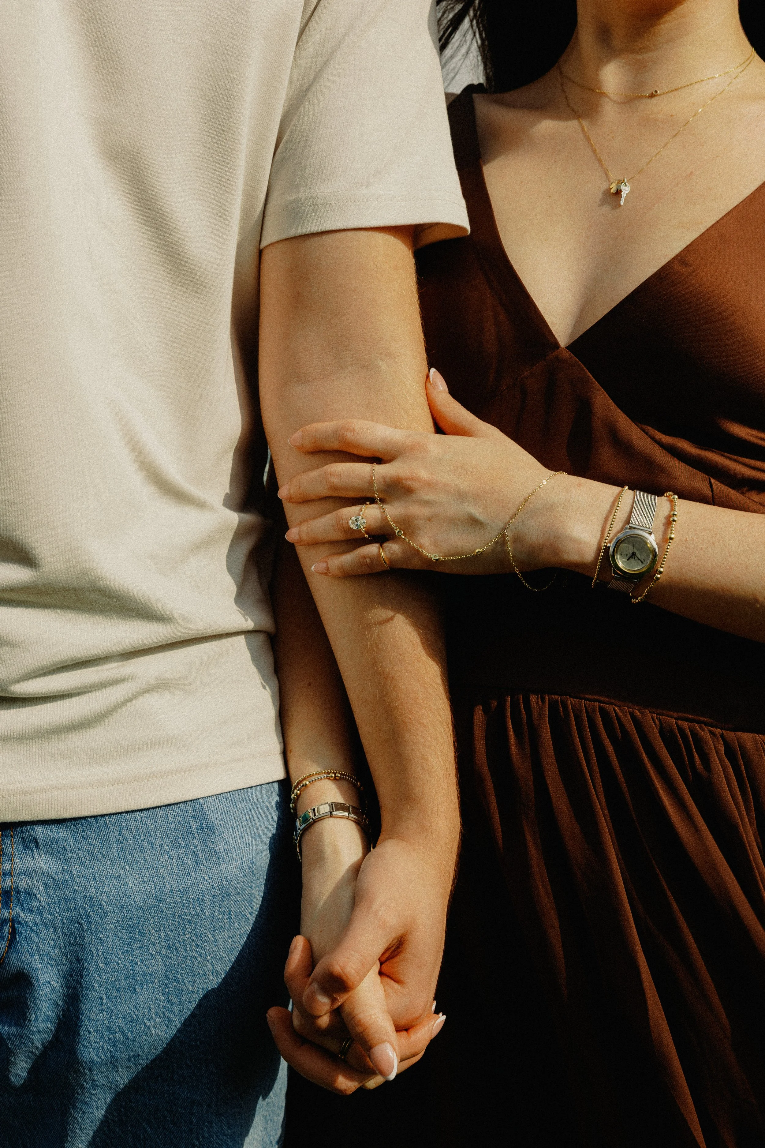 Close-up of two women holding hands, wearing jewelry, with one in a beige T-shirt and jeans, and the other in a dark brown dress.