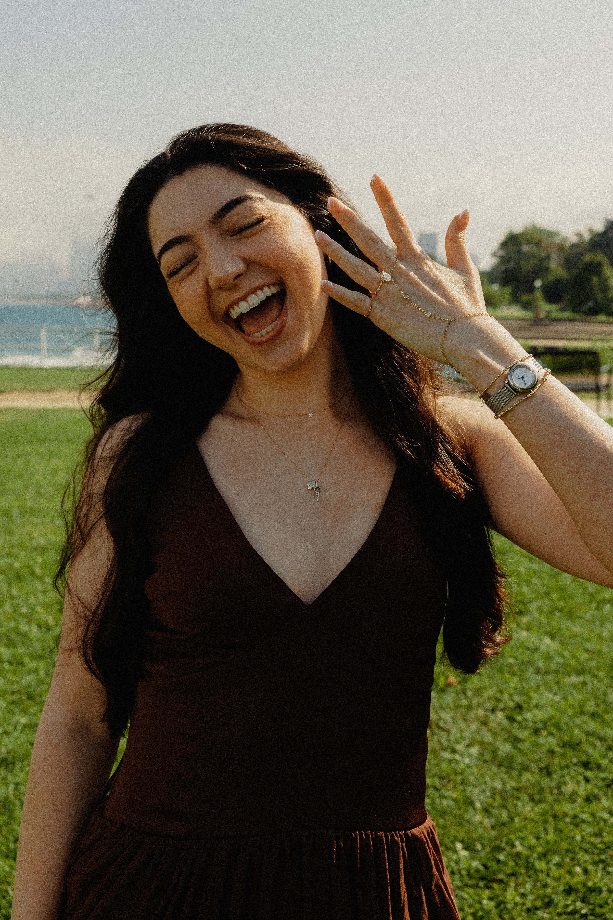 A woman with long dark hair and light skin, laughing outdoors in sunlight, wearing a dark top, with jewelry including rings, a bracelet, a watch, and a necklace, in a park near water with green grass and trees.