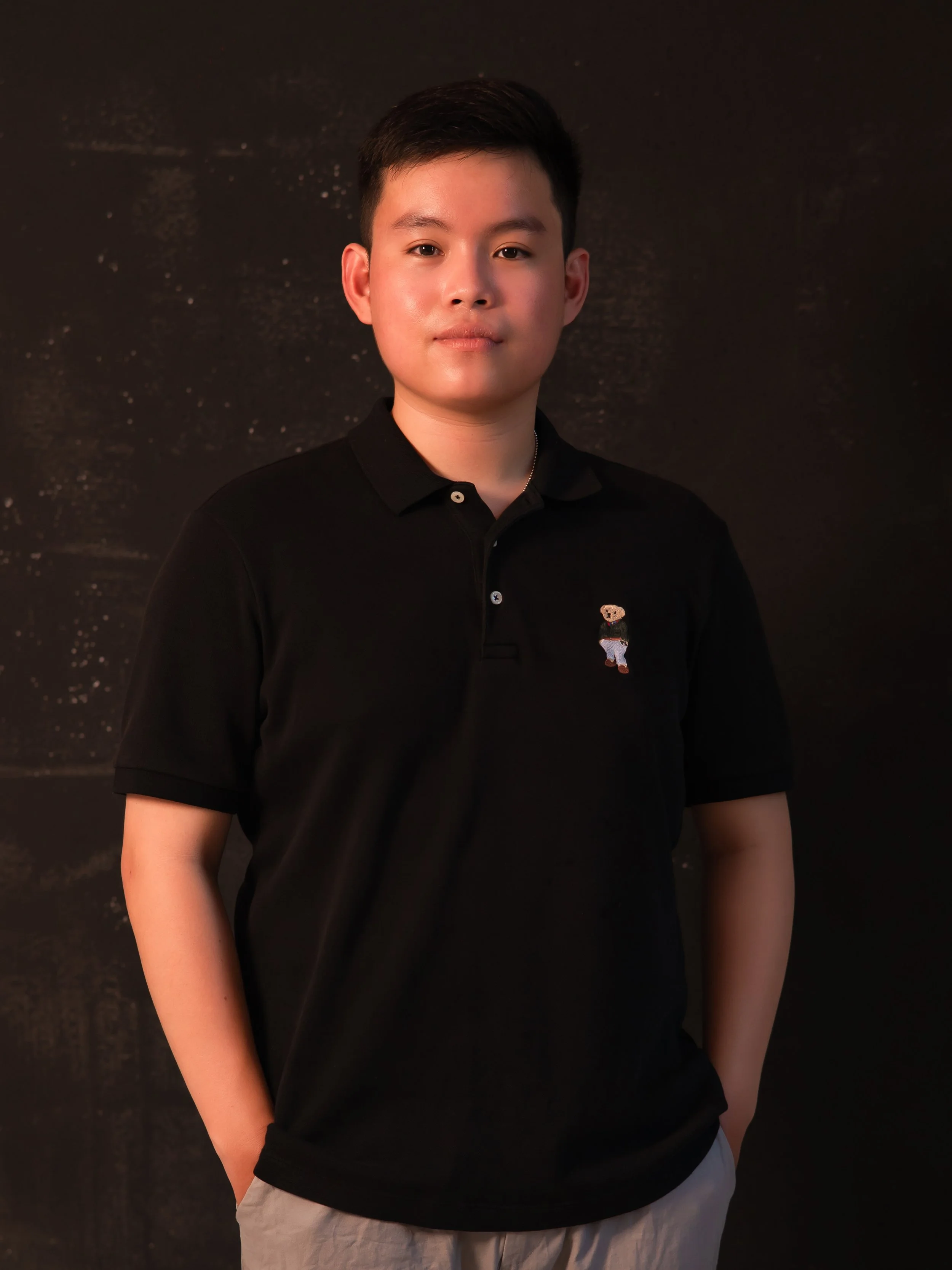 Portrait of a young man with dark hair, wearing a dark jacket over a dark shirt, standing against a plain light-colored background.
