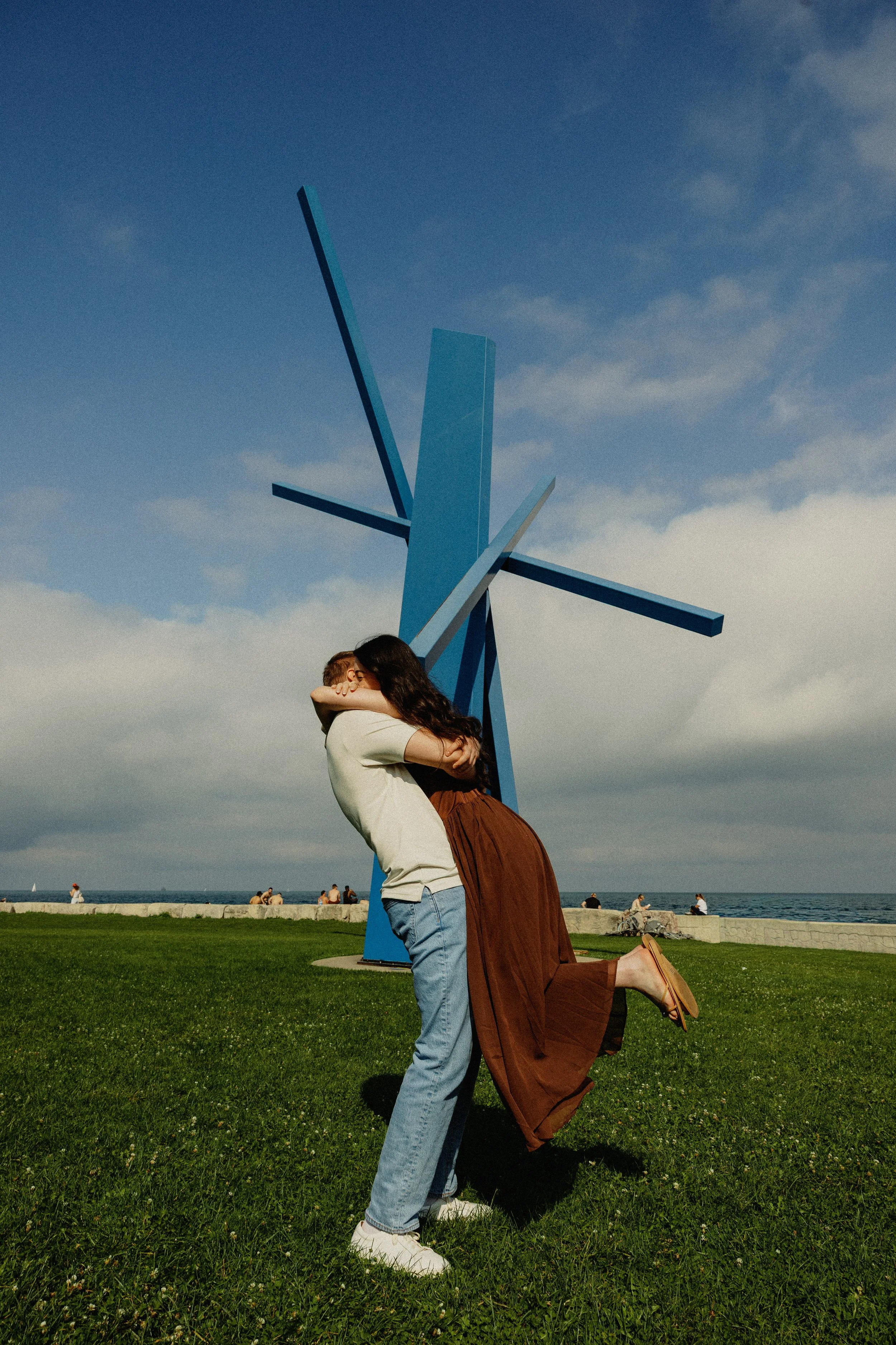 Two people, a man and a woman, embracing by a large blue abstract sculpture on a grassy area near the waterfront on a partly cloudy day. The woman is kneeling with one foot raised, and the man is standing, hugging her.