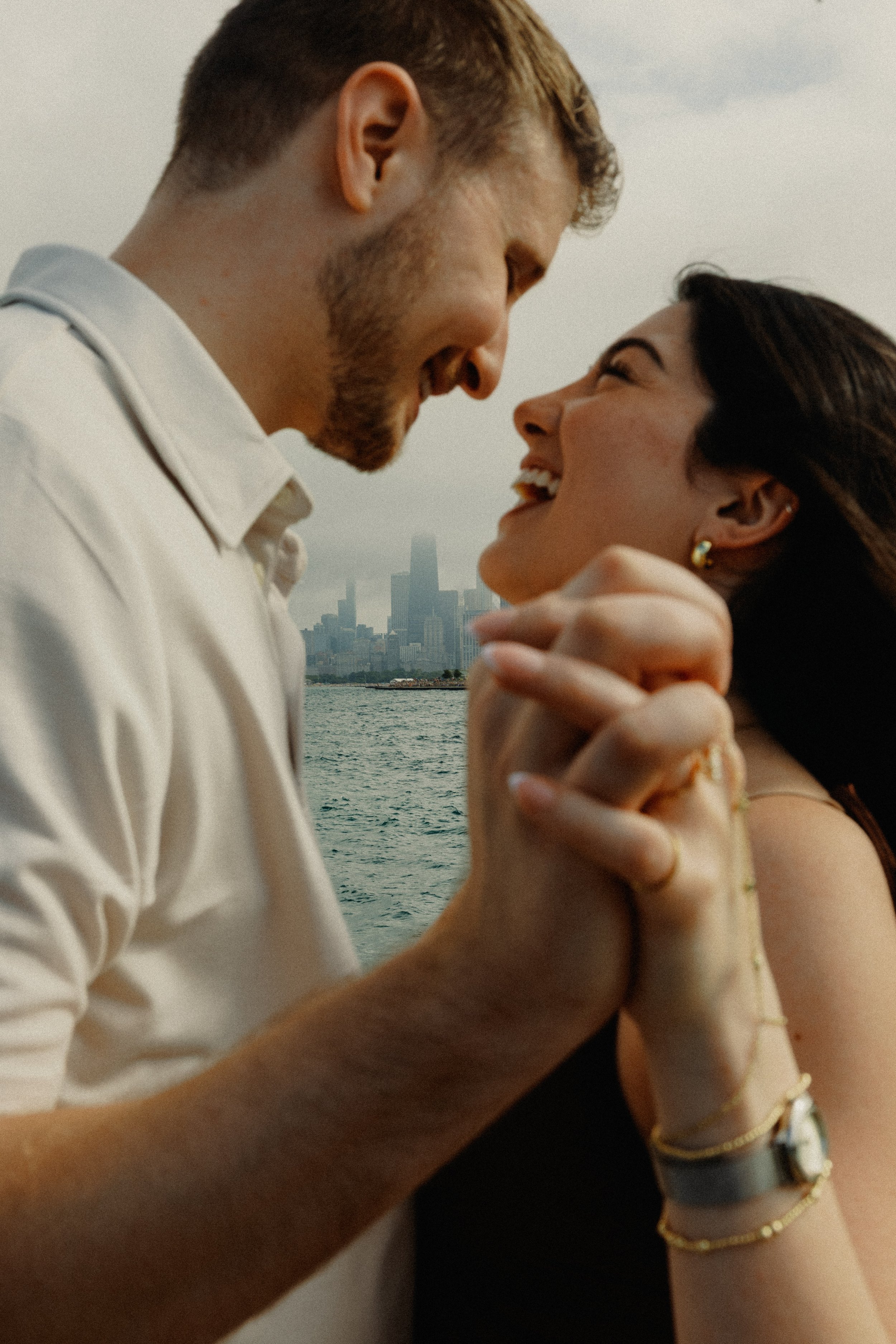 A smiling couple in an affectionate pose near a body of water with a city skyline in the background. The man has short light brown hair and a beard, wearing a white shirt. The woman has long dark hair, earrings, and a sleeveless dress, holding his ha