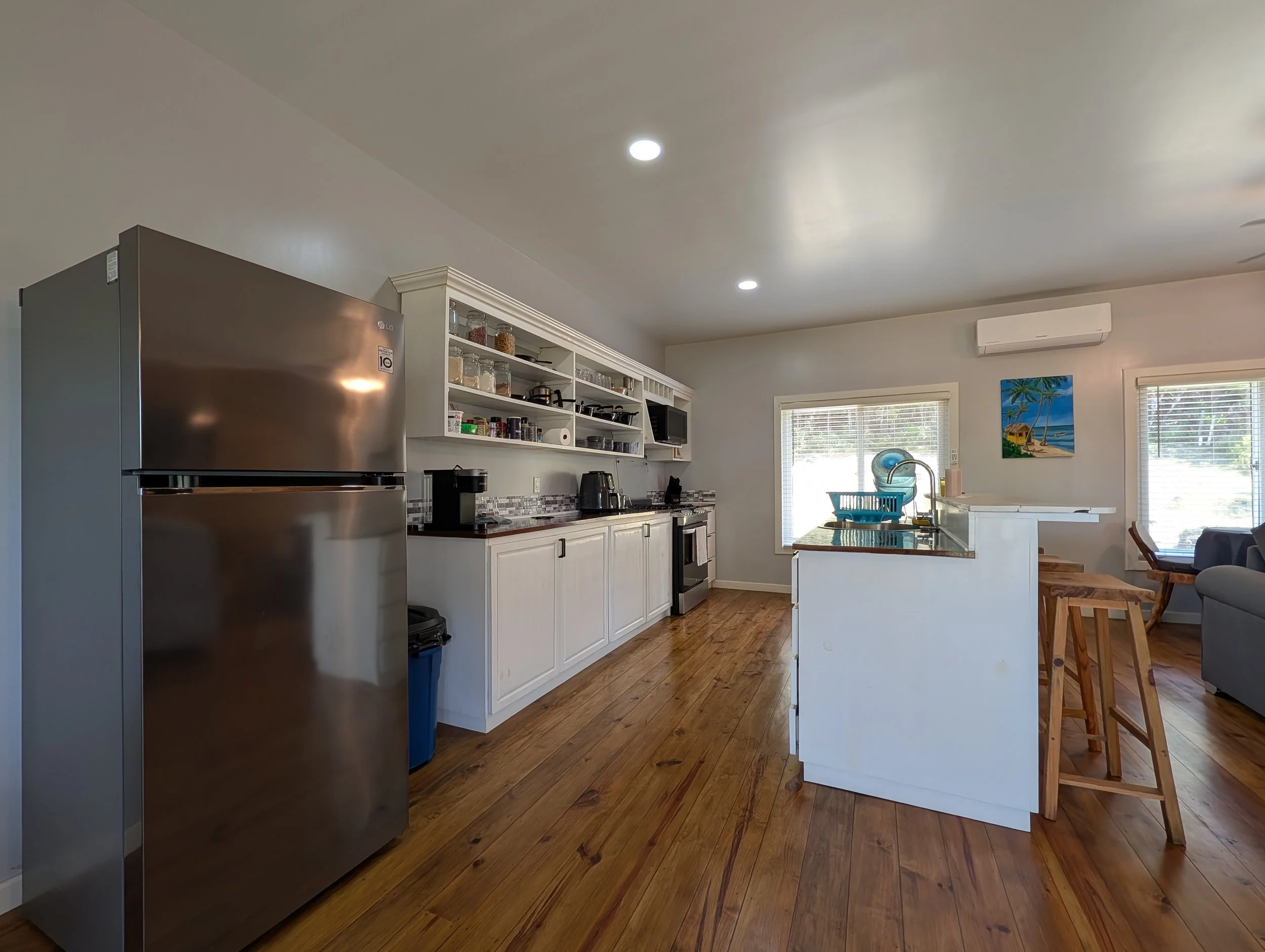 A modern kitchen with white cabinets, open shelving, stainless steel refrigerator, hardwood floors, and a window with blinds. There are small appliances and jars on the counter, and a colorful painting on the wall.