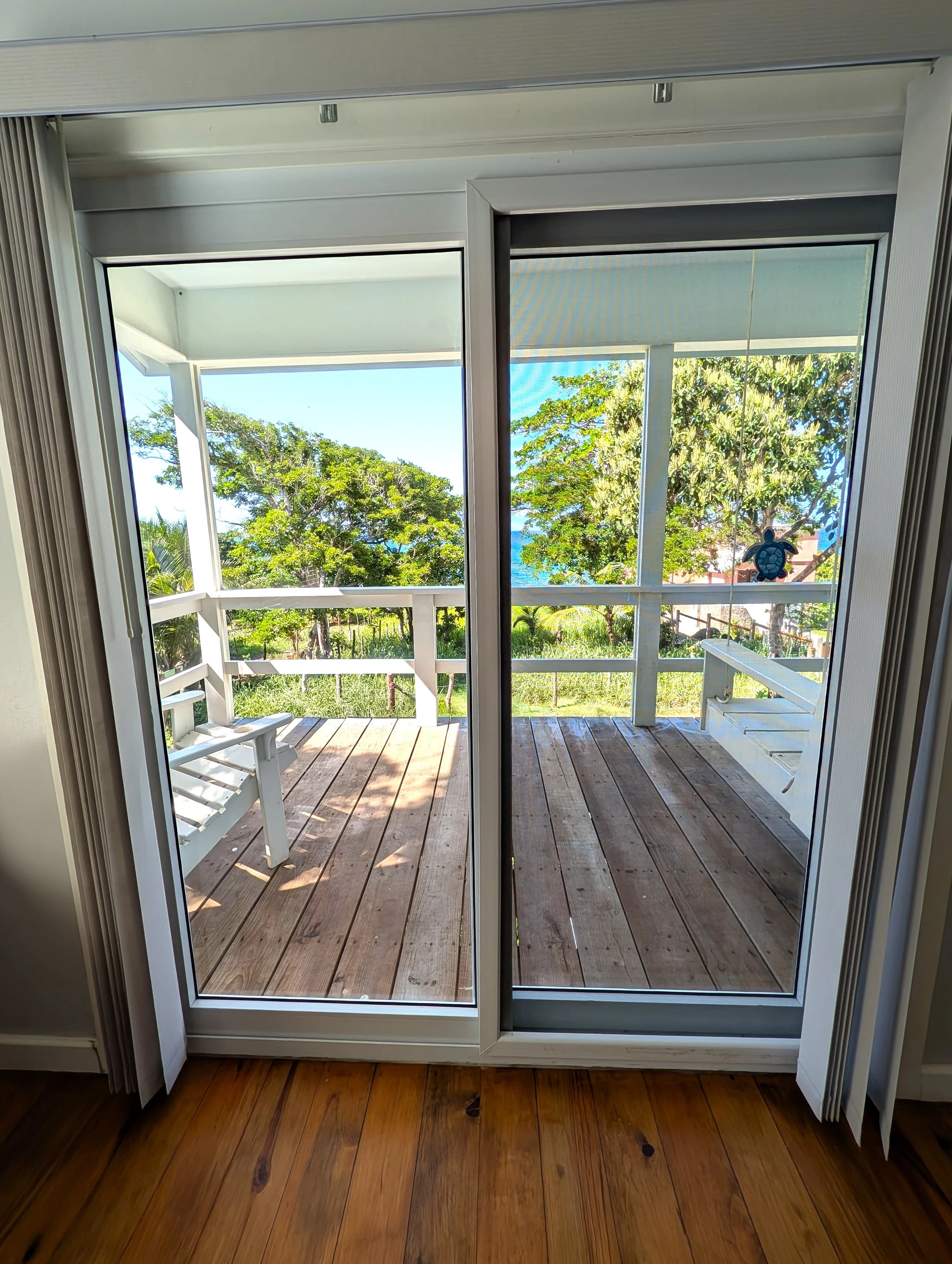 View through sliding glass door onto a wooden porch with trees and greenery outside. The door has a white frame with a turtle wind chime hanging inside. The indoor floor is wooden.