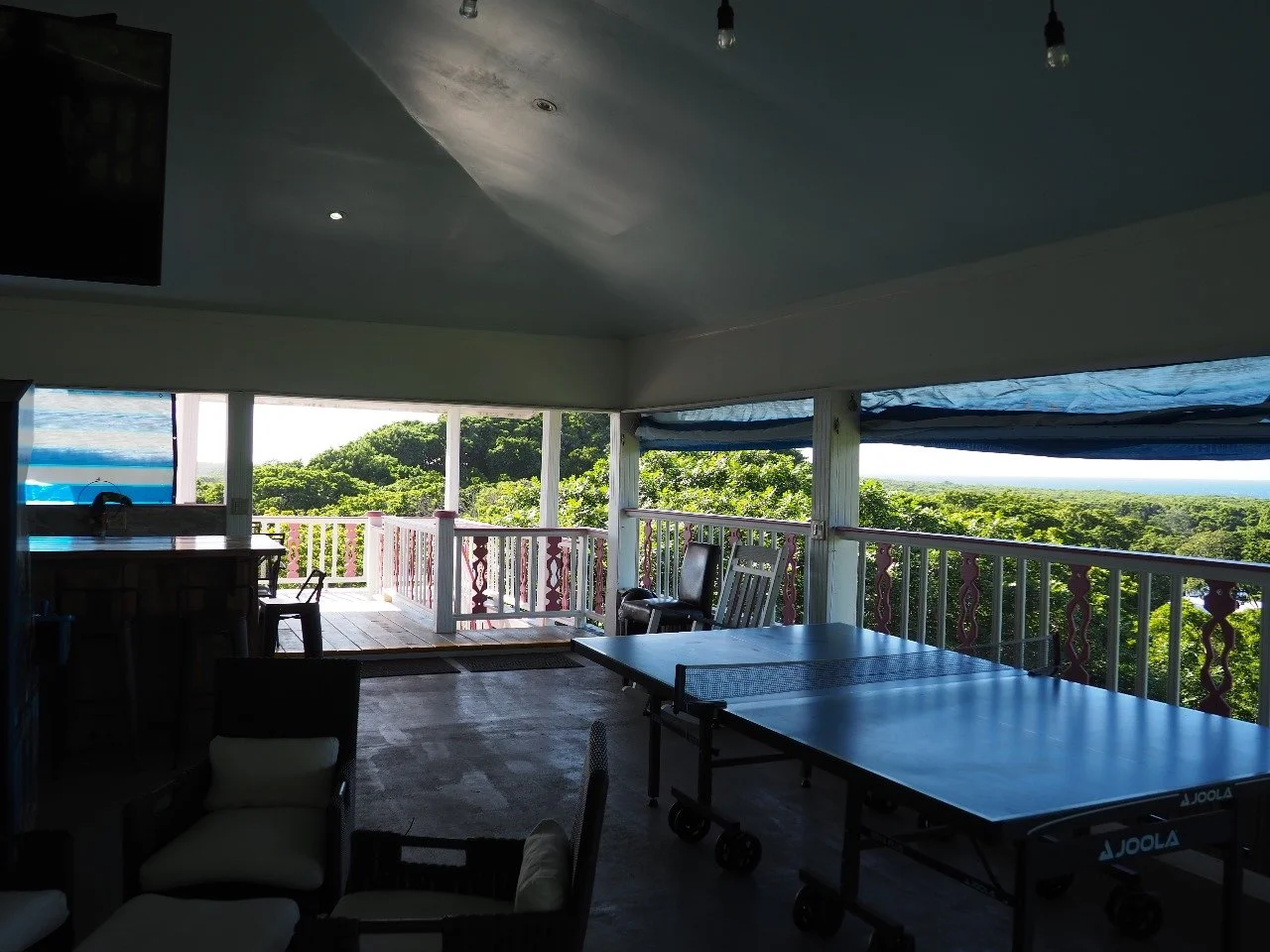 Pink Palace Utila Covered outdoor recreation room with two blue JOOLA ping-pong tables, patio seating, and a lush ocean-view landscape