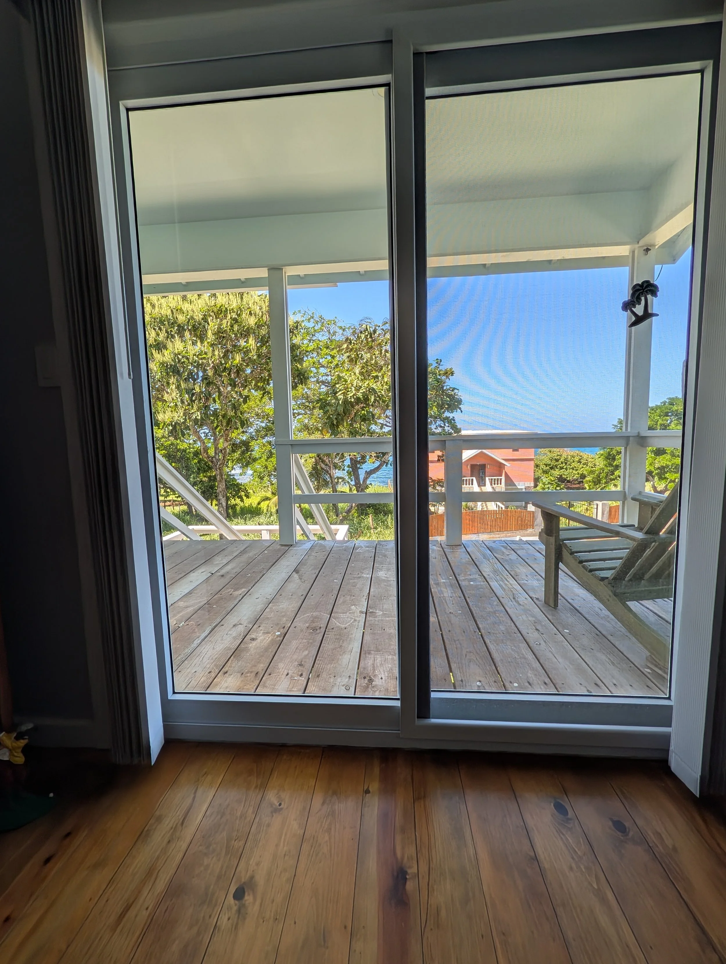 View from inside a house looking out through a sliding glass door onto a wooden porch, trees, blue sky, and neighboring houses in the distance.