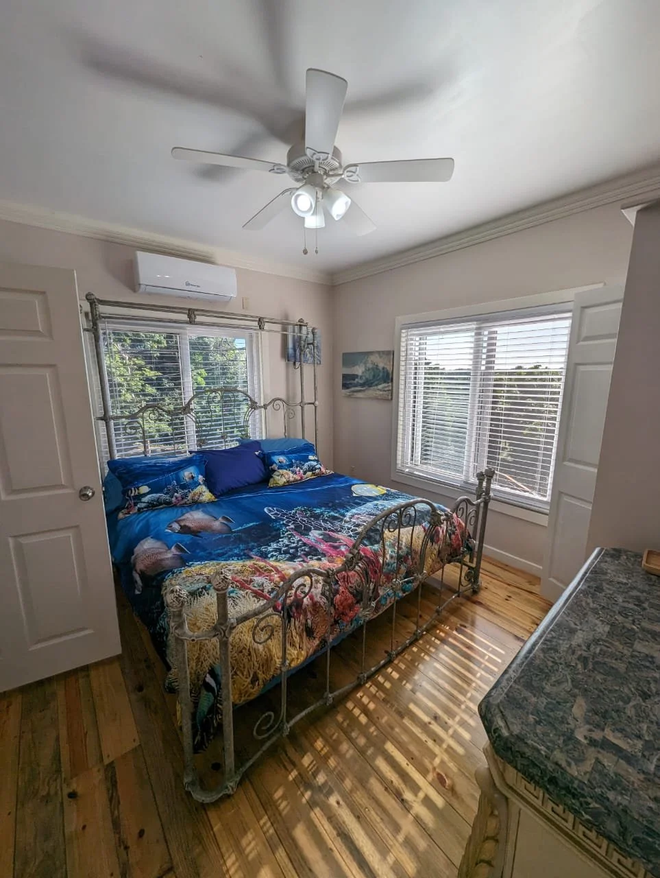 Pink Palace Utila Bright bedroom with a wrought-iron bed, colorful ocean-themed bedding, ceiling fan, windows with blinds, and hardwood floors