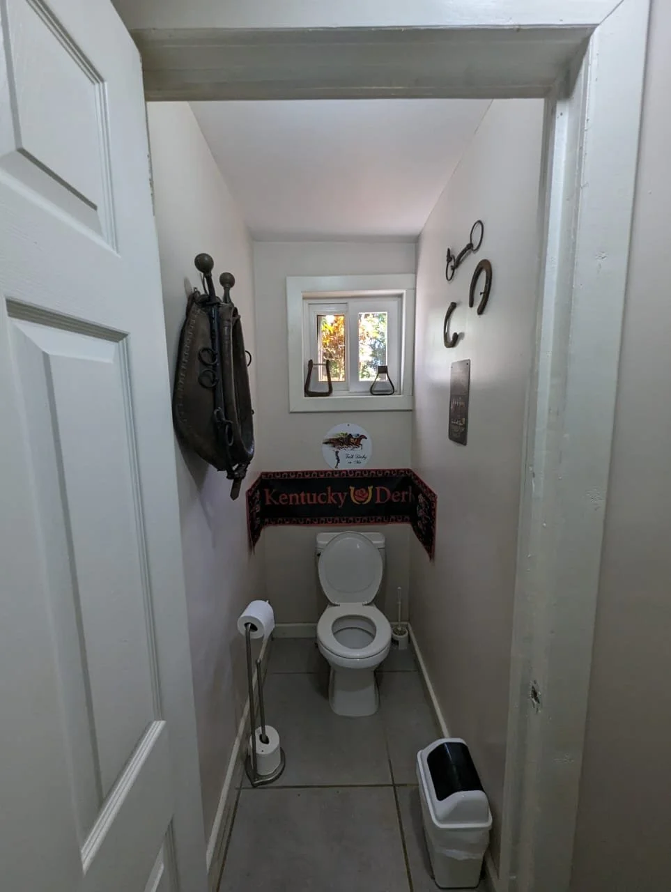 Pink Palace Utila Narrow rustic half-bathroom with a toilet, window, toilet paper holder, wastebasket, and Kentucky Derby wall decor.