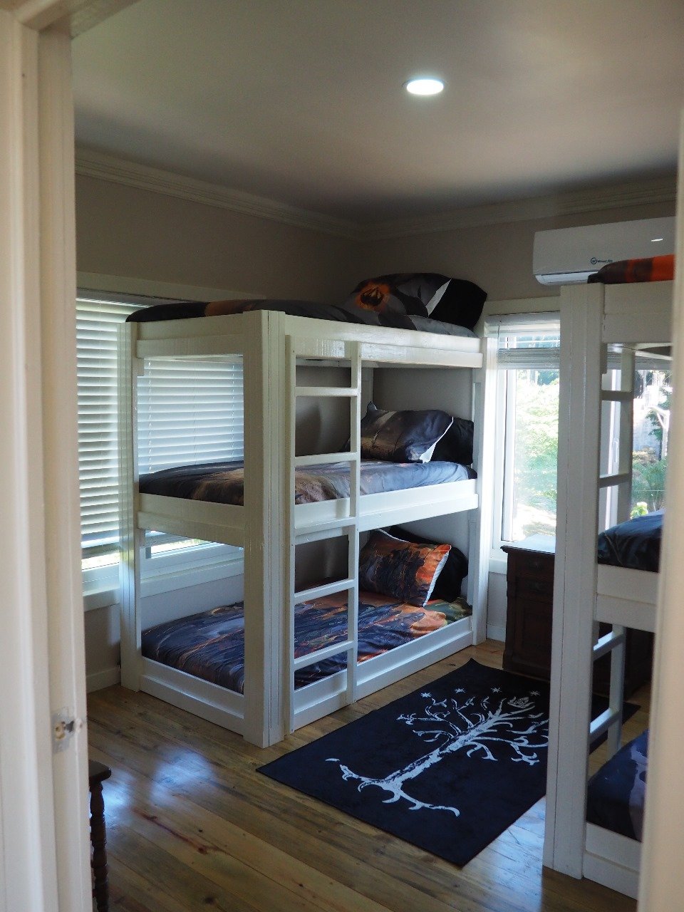 Pink Palace Utila Bright bedroom with white bunk beds, colorful bedding, windows with blinds, air conditioner, and black decorative rug