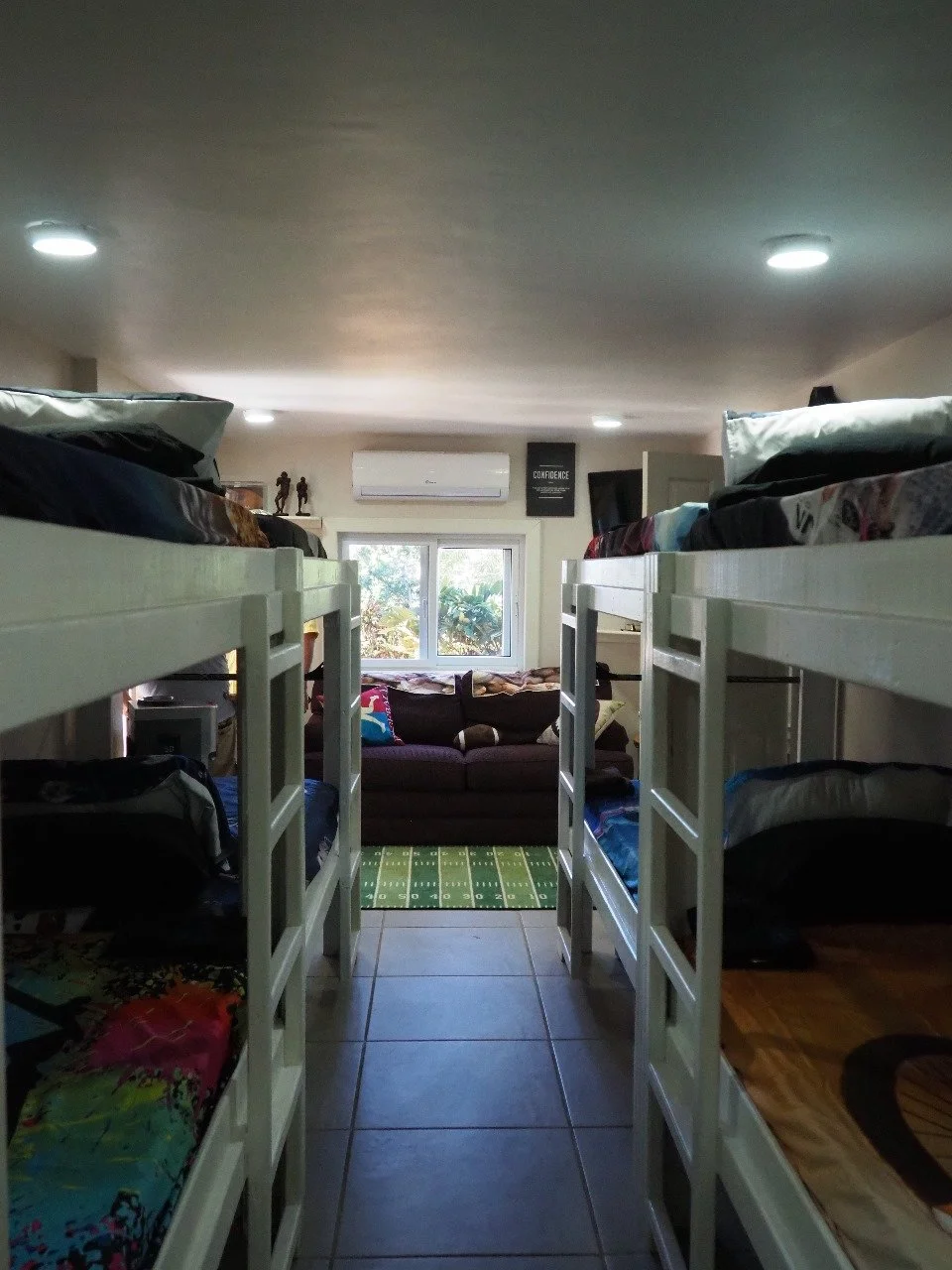 Pink Palace Utila Shared dorm room with white bunk beds, colorful bedding, a couch, window, and wall-mounted air conditioner
