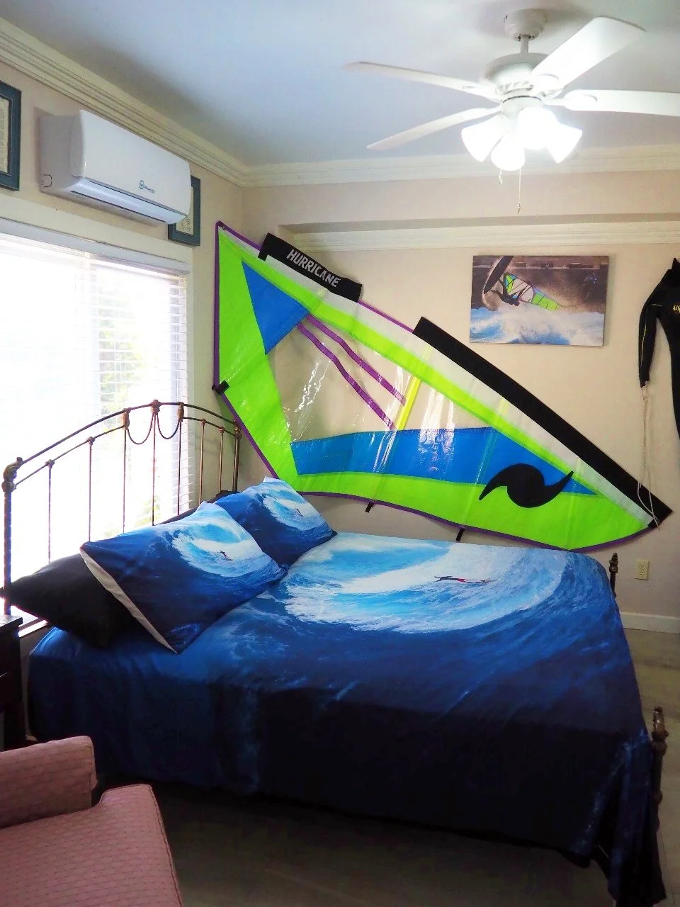 Pink Palace Utila Bright blue ocean-themed bedroom with a bed, colorful windsurfing sail mounted on the wall, ceiling fan, and window air conditioner.