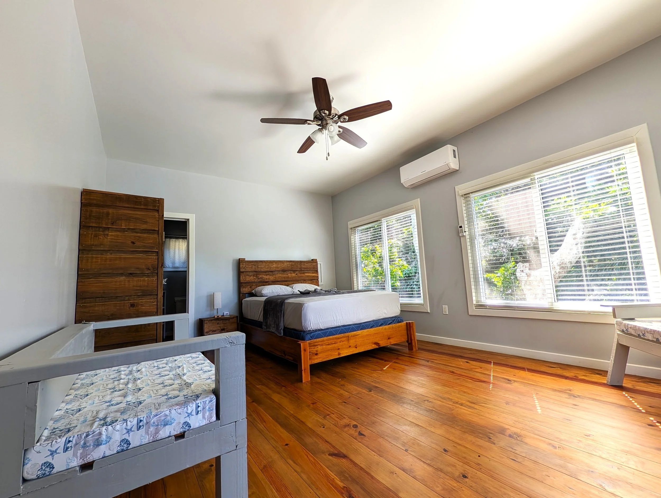 A bright bedroom with wooden floors, white walls, and two large windows with blinds allowing sunlight in. There is a bed with a wooden headboard and a dark gray cover, a small bedside table, a white air conditioning unit on the wall, and a ceiling fan with wooden blades.