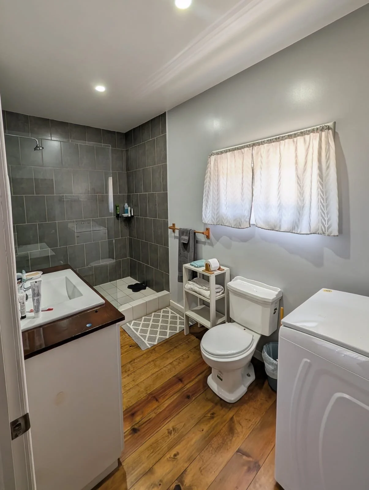 A bathroom with a walk-in shower with gray tiles, a white toilet, a small white shelf with towels and a roll of toilet paper, a washing machine, a white sink with toiletries, a window with white curtains, and wooden flooring.