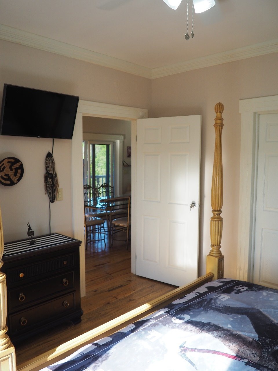 Pink Palace Utila Bedroom with a wall-mounted TV, open door to a dining area, dark dresser, ceiling fan, and wooden bed frame.