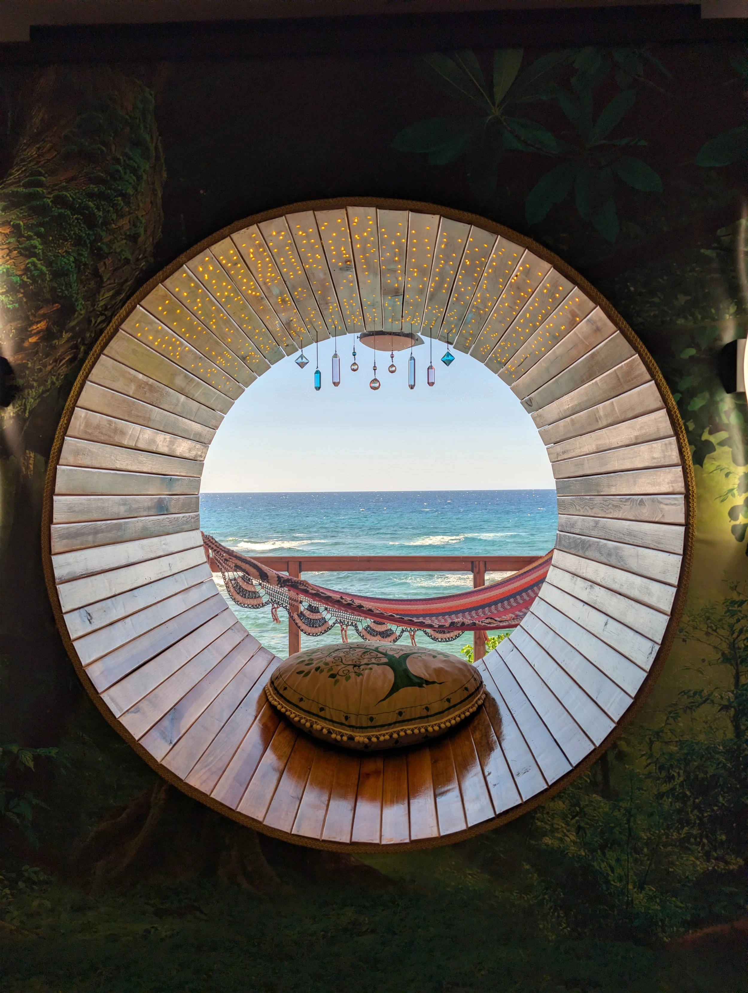 A round window with a wooden frame, decorated with hanging colorful pendants, opens to a view of the ocean with waves, a clear sky, and a hammock on a balcony.