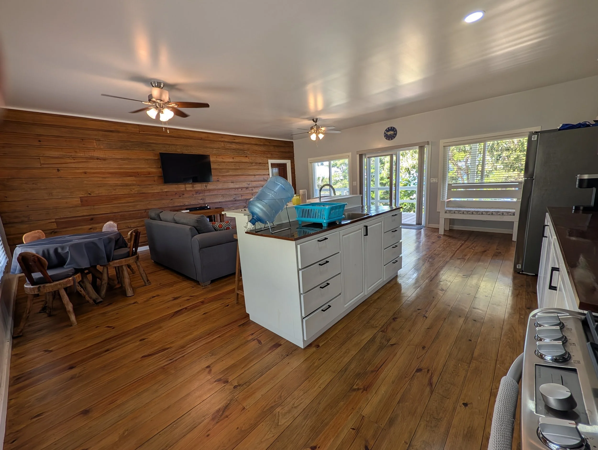 Open kitchen and living room with wooden flooring, white cabinets, a stainless steel refrigerator, a television mounted on a wood-paneled wall, and sliding glass doors leading outside with trees visible.