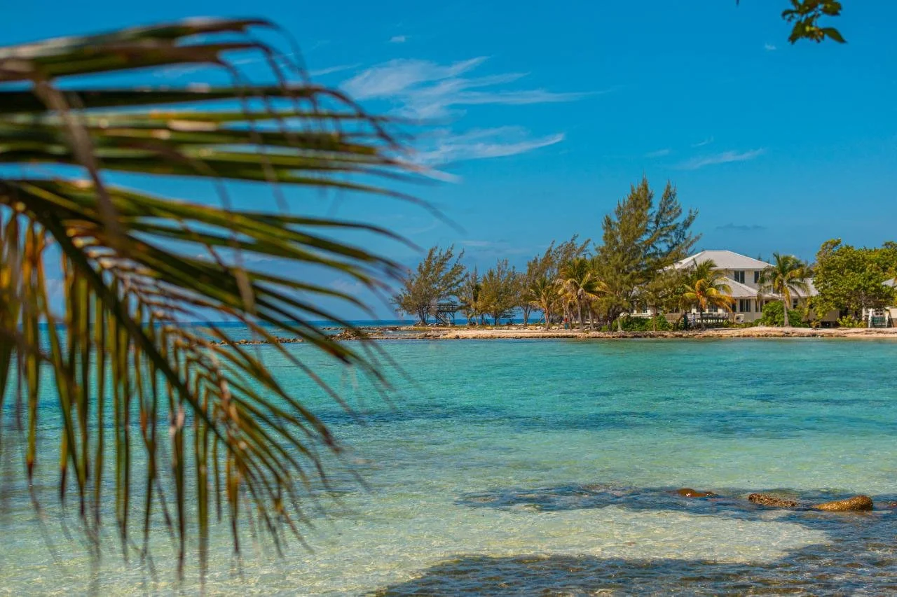 Tropical beach with turquoise waters, sandy shoreline, palm trees, and a white house in the background under a blue sky.