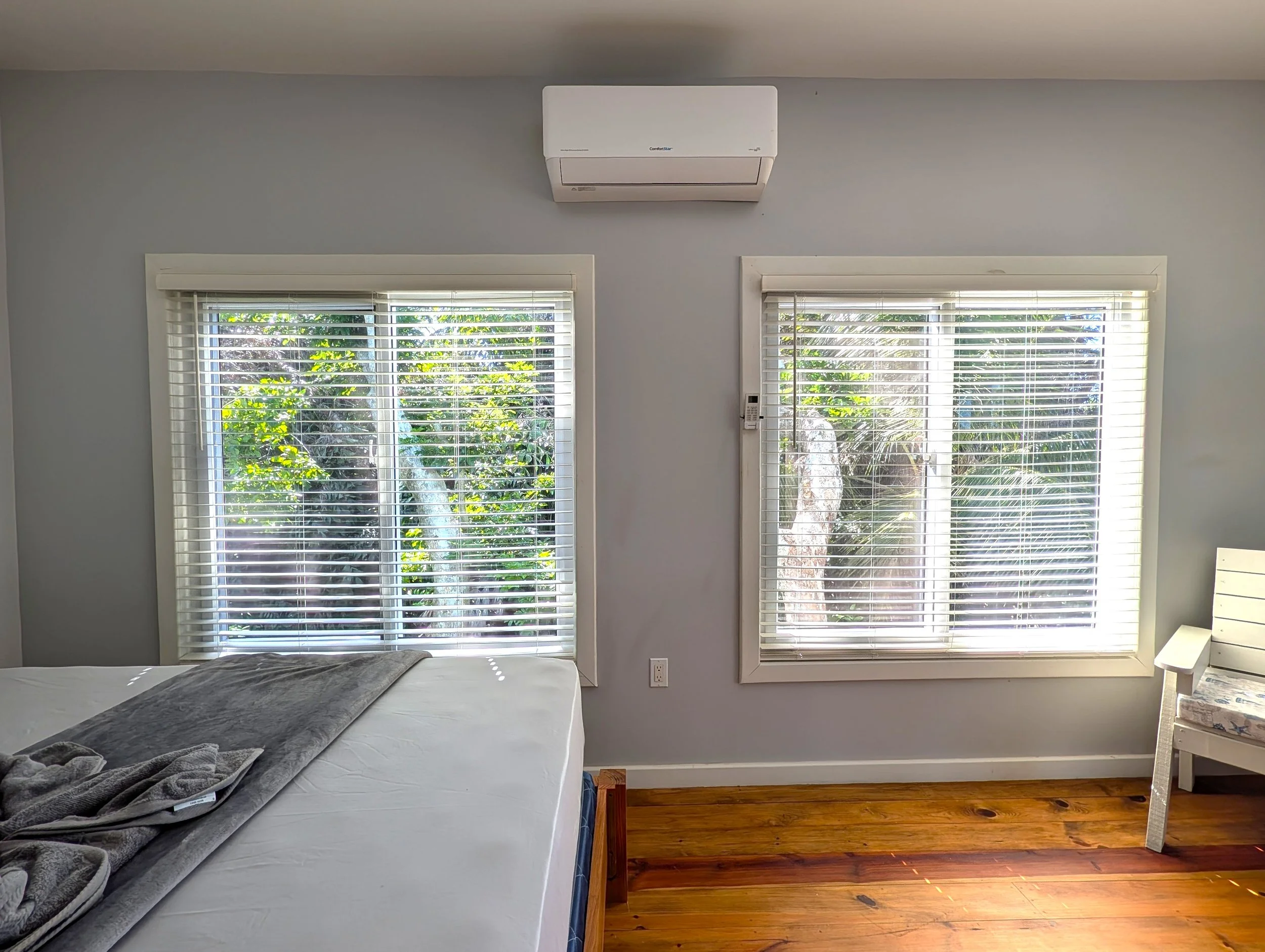 A bedroom with two large windows with white blinds showing greenery outside. A white bed with a grey blanket and some clothes on it is positioned in front of the left window. To the right of the windows, a wall-mounted air conditioning unit is visible near the ceiling. A bedroom remote control is attached to the wall between the windows. The room has wooden flooring, and a small part of a white chair is visible in the bottom right corner.
