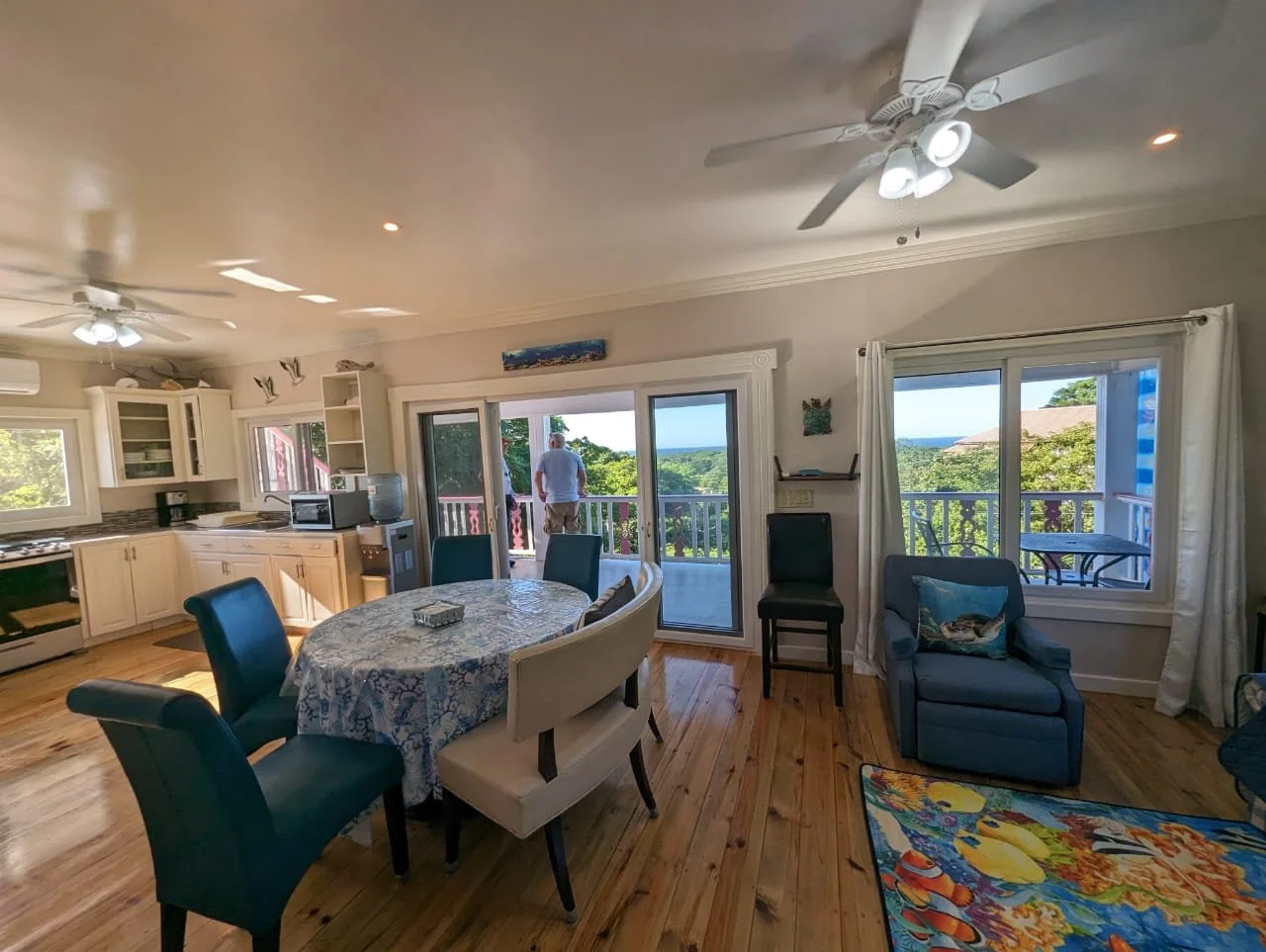 Pink Palace Utila Bright coastal vacation home dining area with kitchen, ceiling fans, sliding doors, ocean views, and table surrounded by chairs.