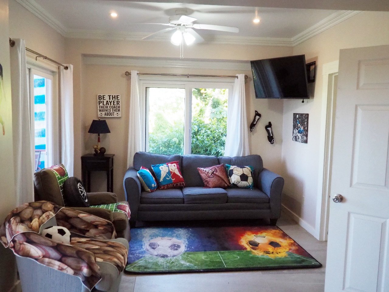 Pink Palace Utila Sports-themed living room with gray sofa, soccer ball pillows, wall-mounted TV, and soccer field rug