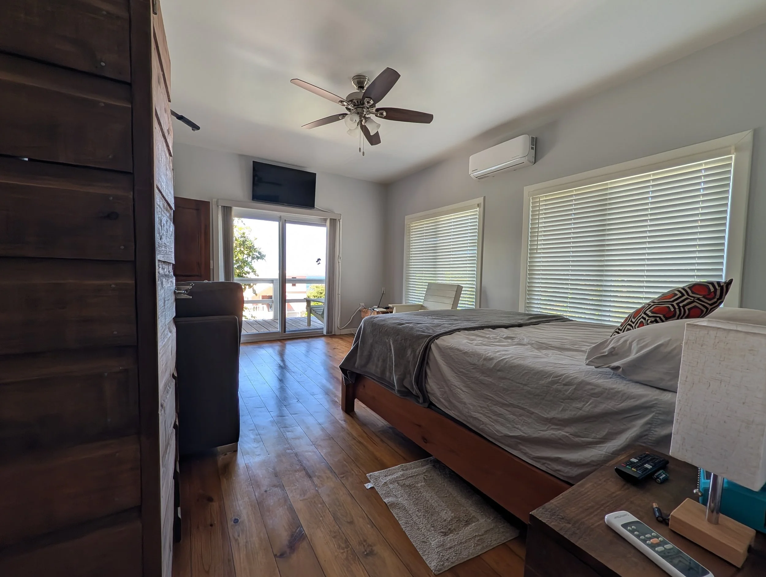 A bedroom with a bed, window blinds, and a sliding glass door leading to a balcony. The room has wooden flooring, a ceiling fan, and an air conditioning unit. There is a remote on the side table and a TV mounted on the wall.