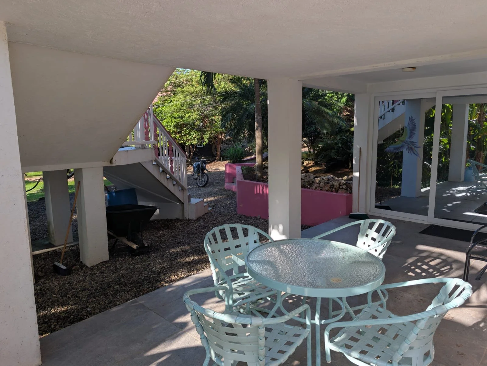 Pink Palace Utila Covered outdoor patio with glass-top table, white wicker chairs, pink planters, tropical garden, and exterior stairs
