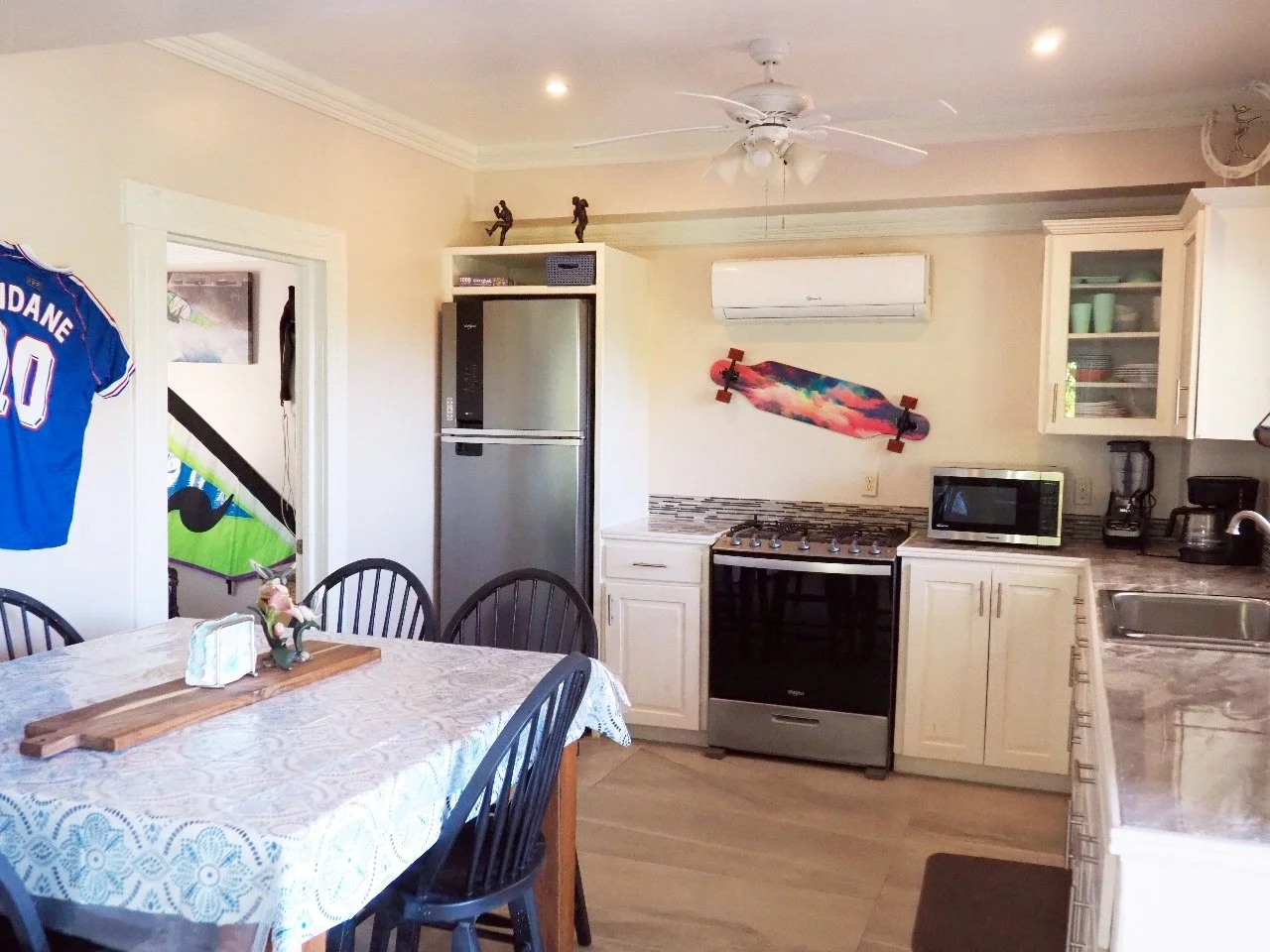 Pink Palace Utila Bright home kitchen with dining table, stainless steel refrigerator, black range, microwave, sink, cabinets, wall-mounted air conditioner, and colorful skateboard decor