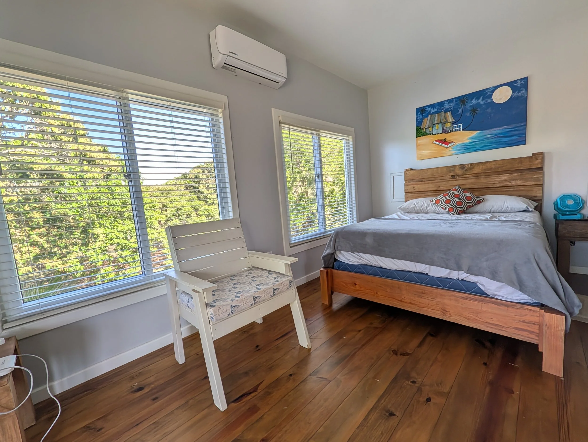 A bedroom with large windows, a bed with a wooden headboard, a colorful painting of a beach scene, a white bench with a patterned cushion, and a small blue side table. Wooden floor and an air conditioning unit on the wall.