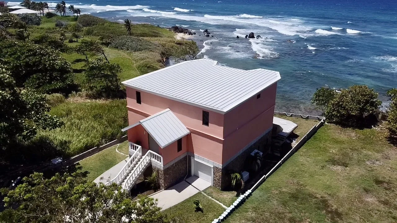A pink house with a white metal roof next to the ocean, surrounded by greenery and trees.