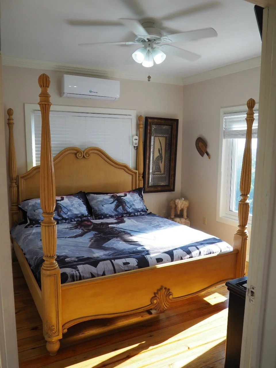 Pink Palace Utila Bright bedroom with an ornate wooden four-poster bed, patterned bedding, ceiling fan, wall-mounted air conditioner, windows, and framed decor.