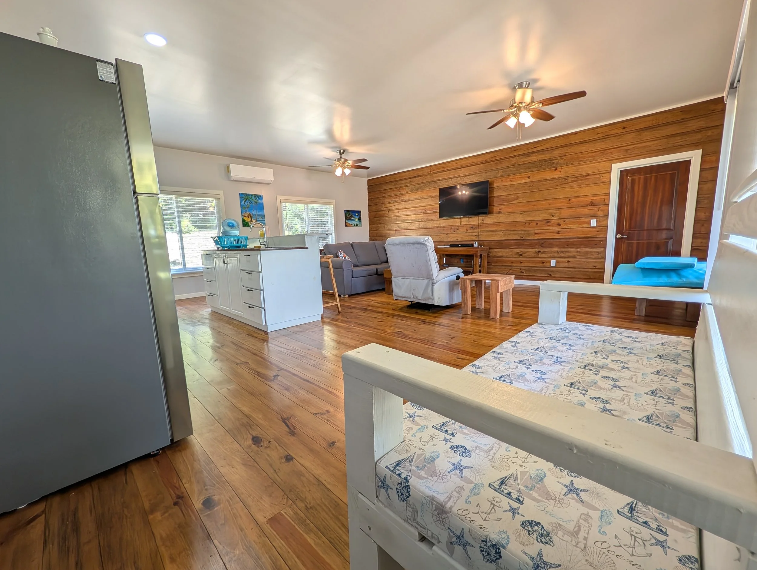 Living room with wood-paneled accent wall, ceiling fans, gray sofa, white armchair, wooden tables, large windows, kitchen with island, refrigerator, and some wall art.