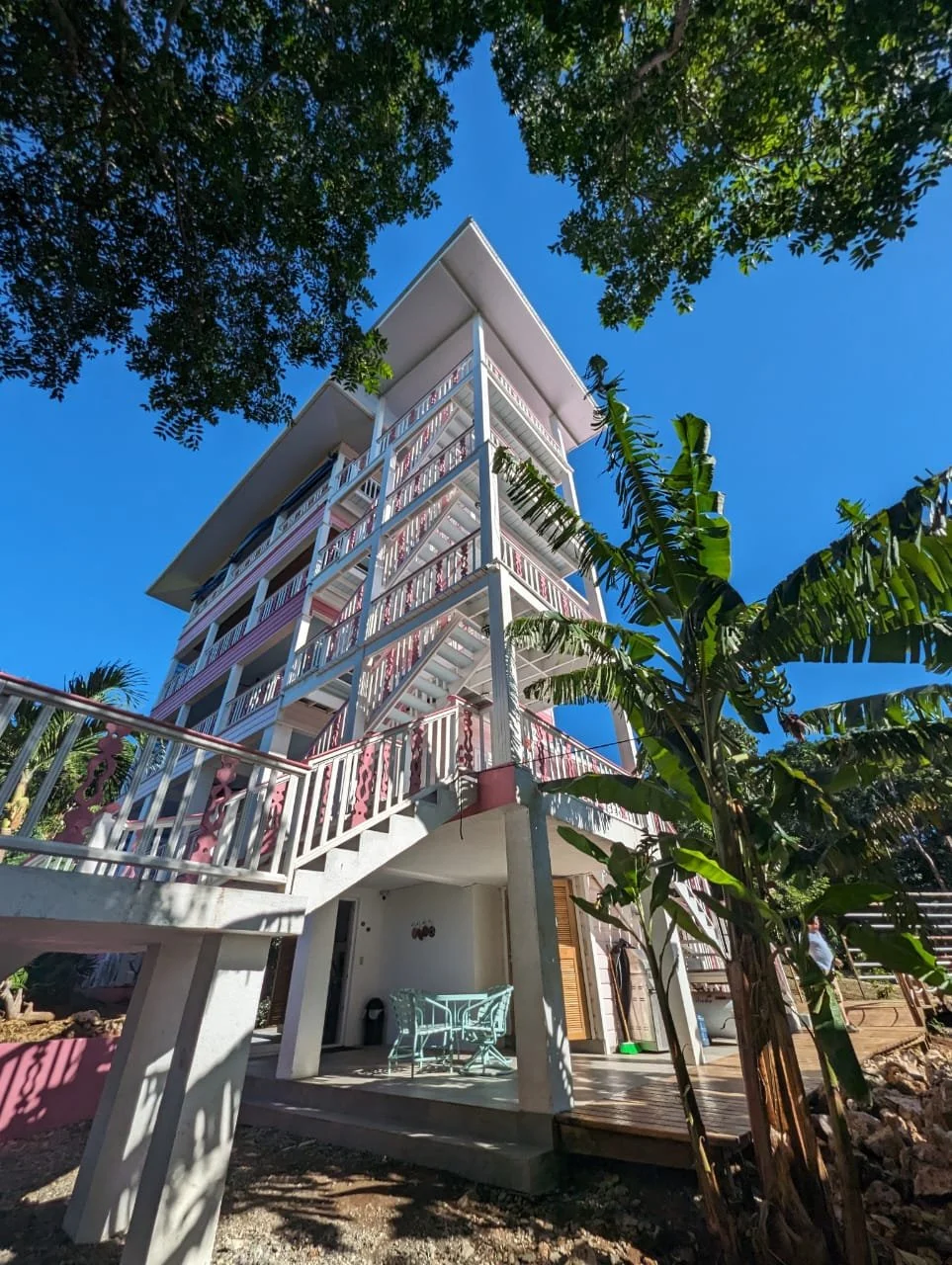 Pink Palace Utila Multi-story tropical building with pink-and-white balconies surrounded by lush palm trees under a bright blue sky