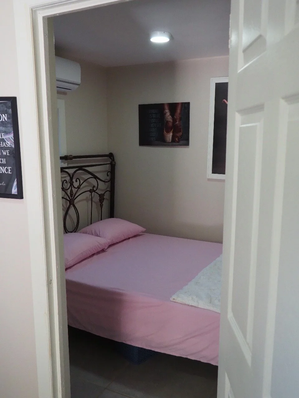 Pink Palace Utila Small bedroom viewed through an open door, featuring a pink-covered bed, ornate black metal headboard, wall art, and a ceiling light.