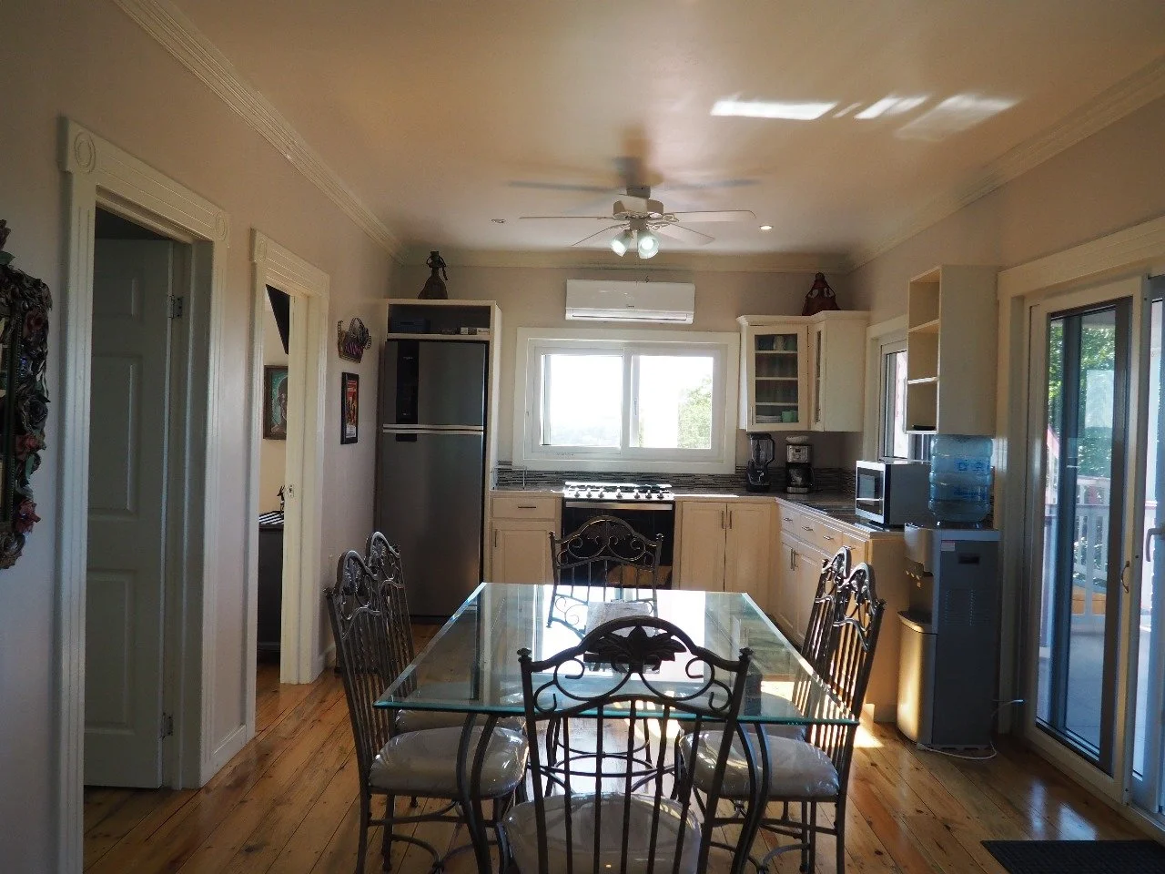 Pink Palace Utila Bright kitchen and dining area with a glass table, wrought-iron chairs, stainless steel refrigerator, stove, cabinets, ceiling fan, and sliding glass doors.