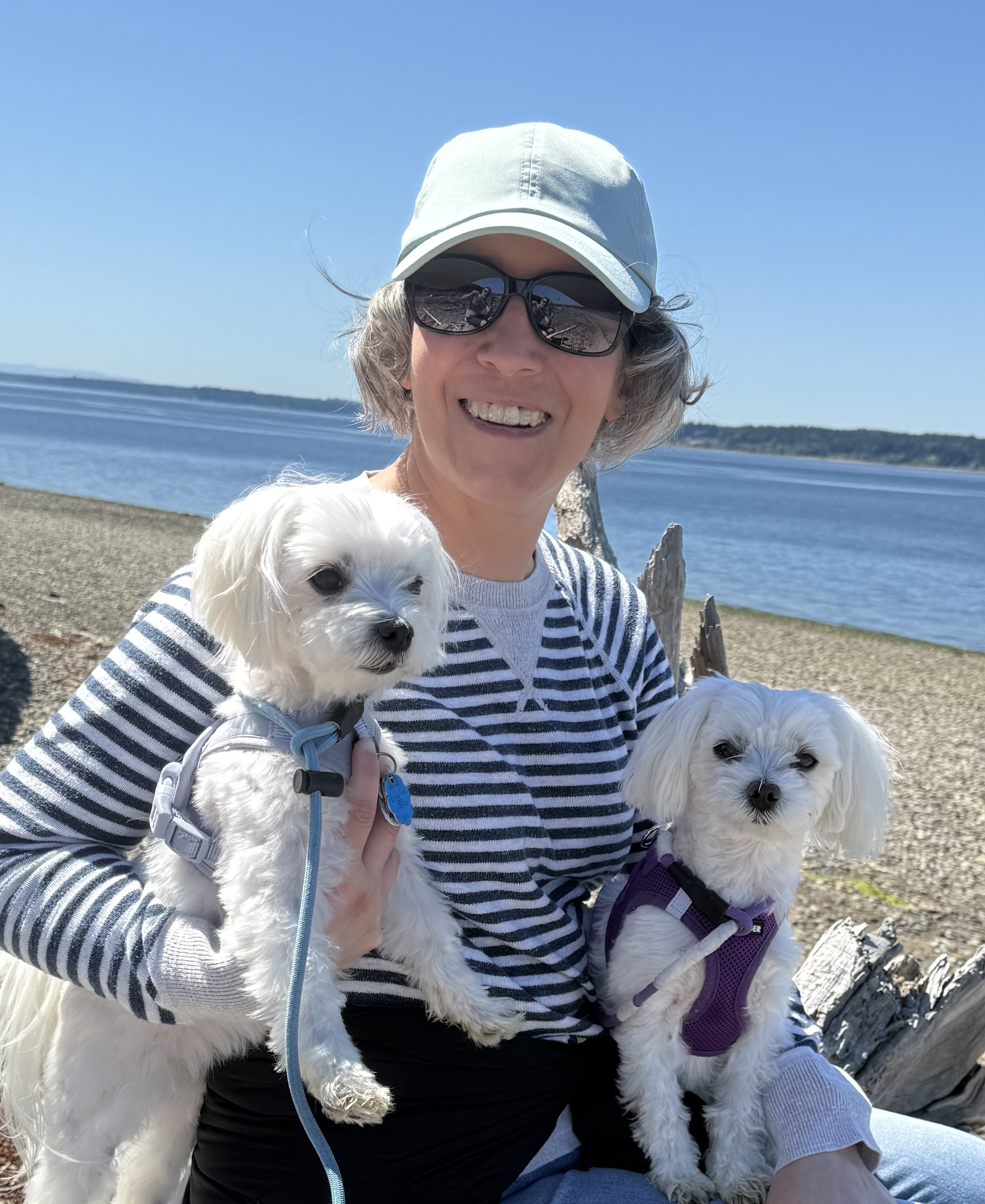 A person smiling at the camera, wearing sunglasses and a light-colored cap, holding two small white dogs, with a beach and water in the background on a sunny day.