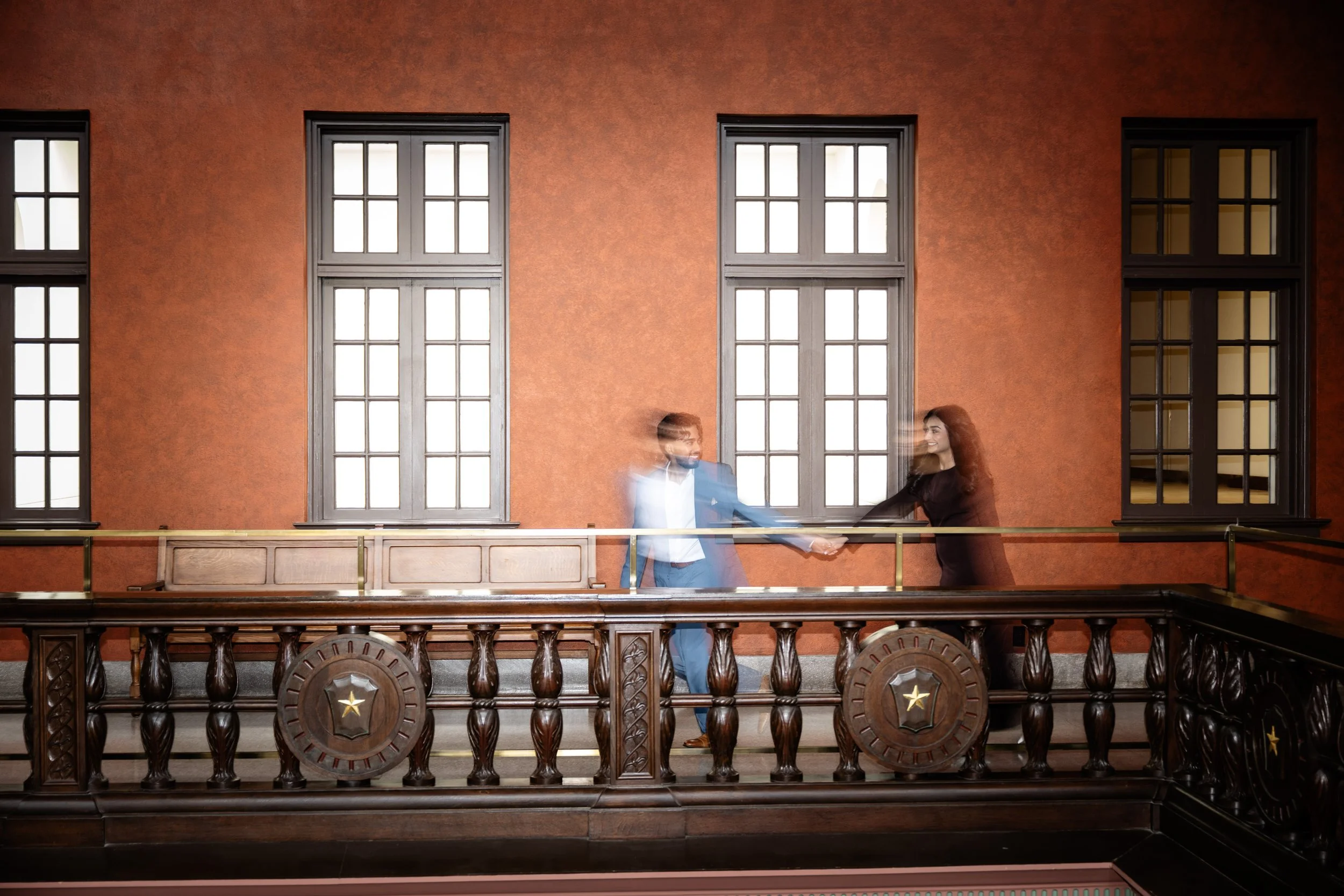 A man in a blue suit and a woman in a black dress are holding hands and dancing on a wooden balcony with a dark wooden railing, in front of a red brick wall with three tall windows with black frames.