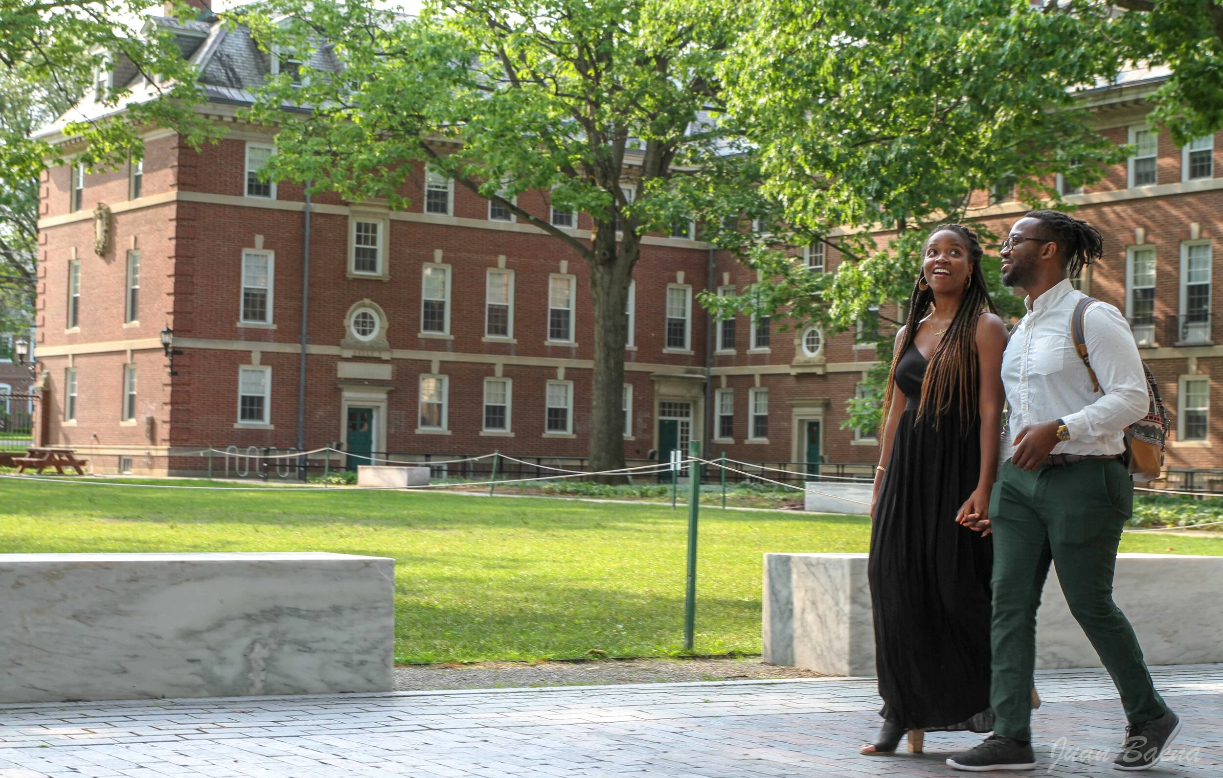 A couple walking together outdoors on a sunny day, with green trees and a red brick building in the background.