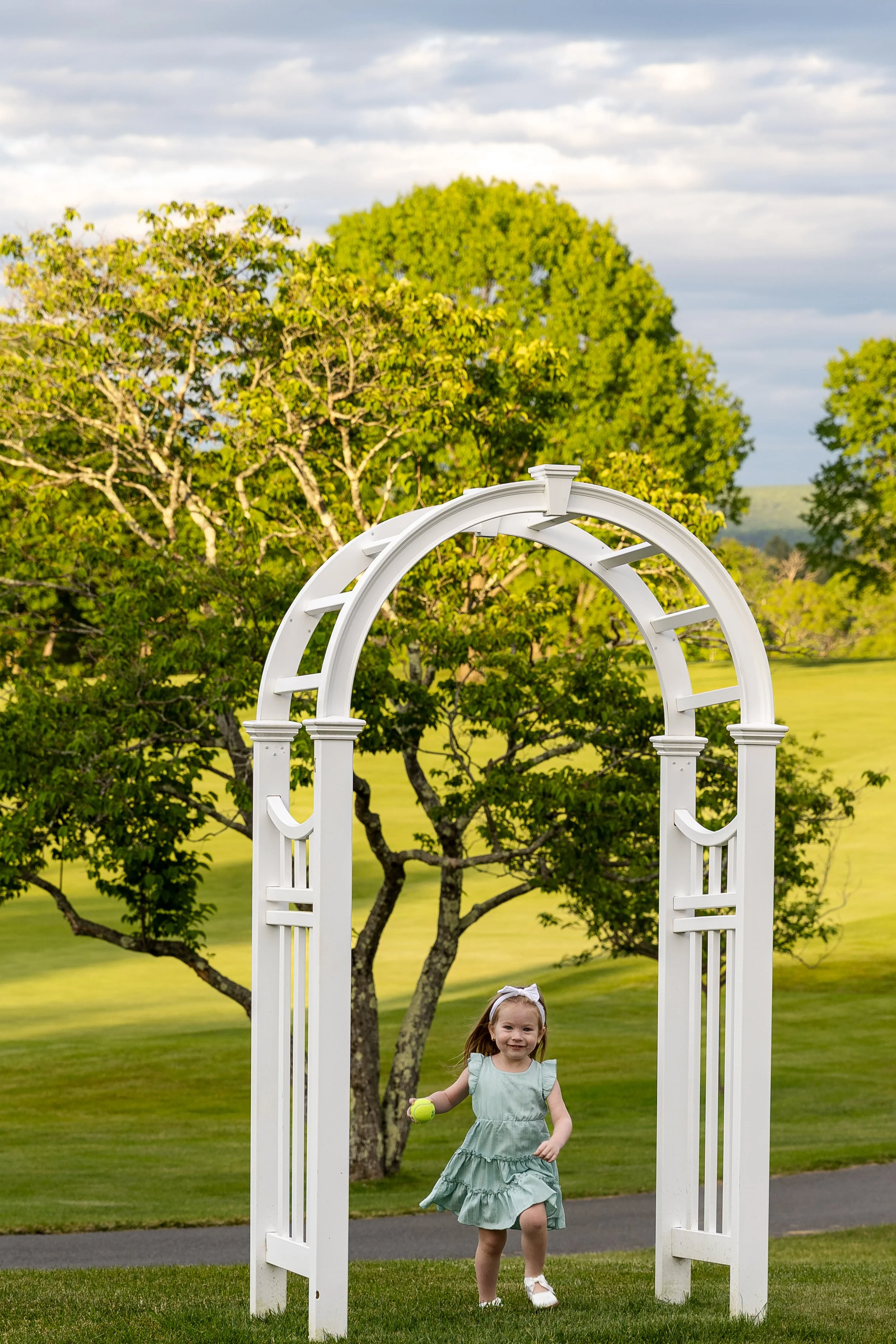 A young girl in a light green dress with ruffles and white shoes is running on a grassy area, holding a tennis ball, beneath a white garden arch, with trees and a cloudy sky in the background.