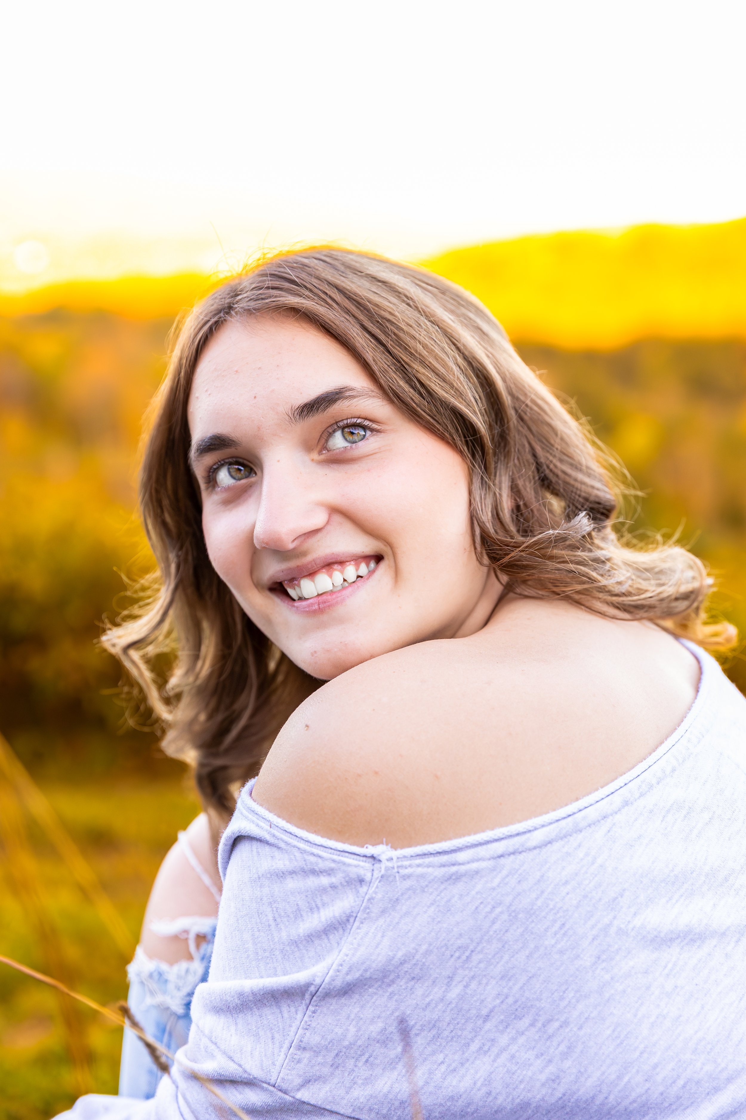 A young woman with light brown wavy hair and light skin smiling outdoors during sunset, with a blurred background of yellow foliage.