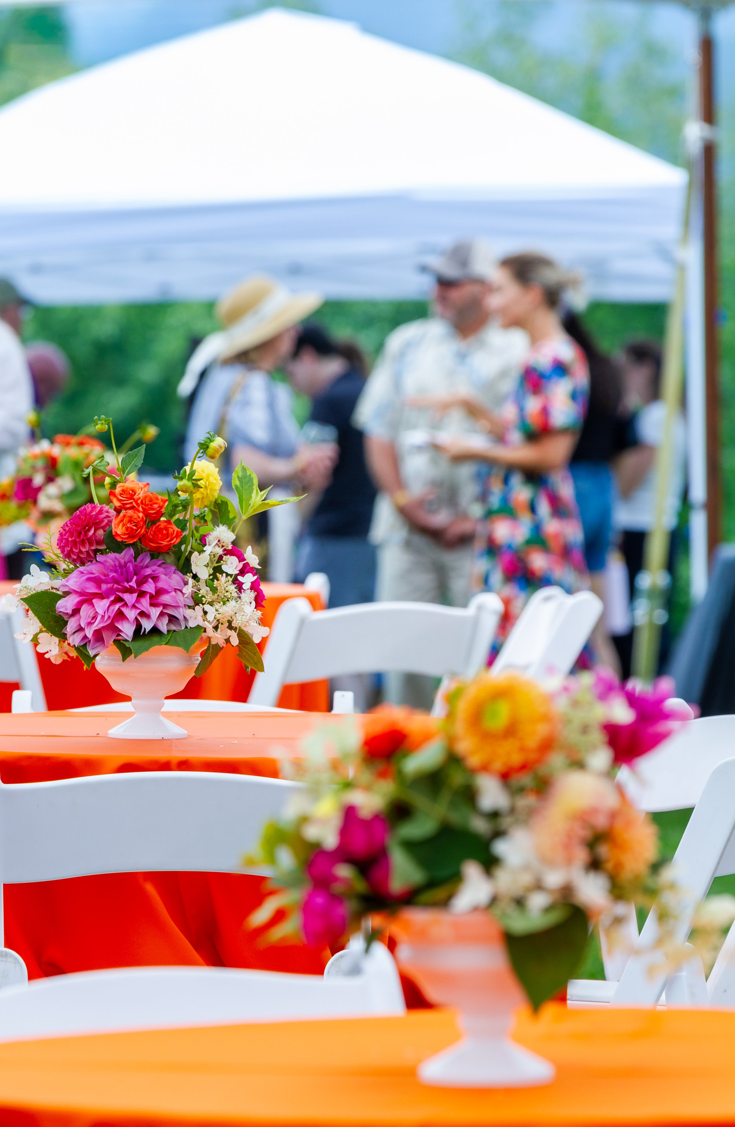 Colorful flower arrangements on orange tables at an outdoor gathering with blurred people in the background under a white canopy.