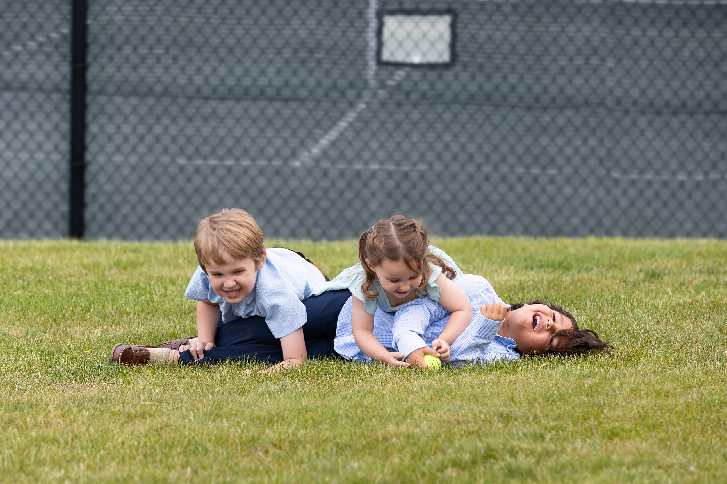 Three children playing on the grass during the day, with a chain-link fence in the background. The children are smiling and appear to be having fun.