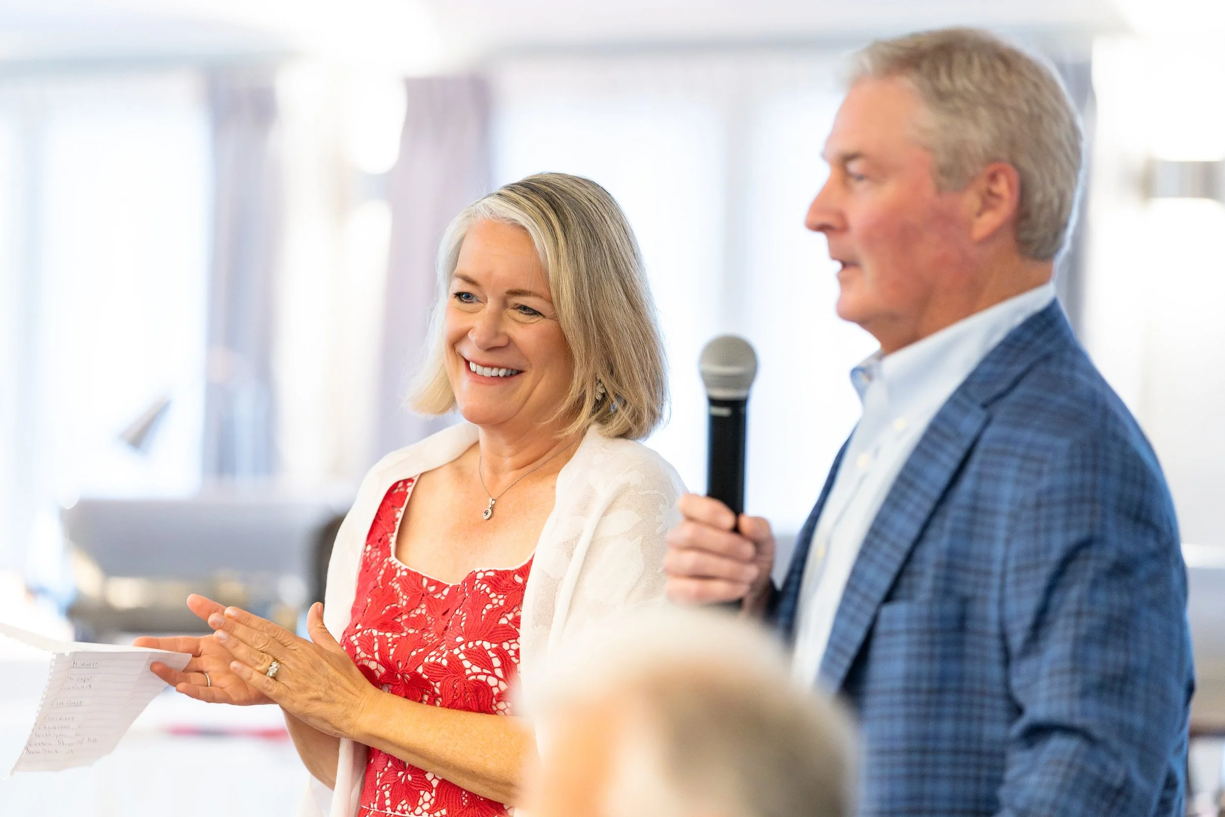 A smiling woman in a red dress with lace details listens as a man in a blue plaid suit speaks into a microphone during a presentation or speech.