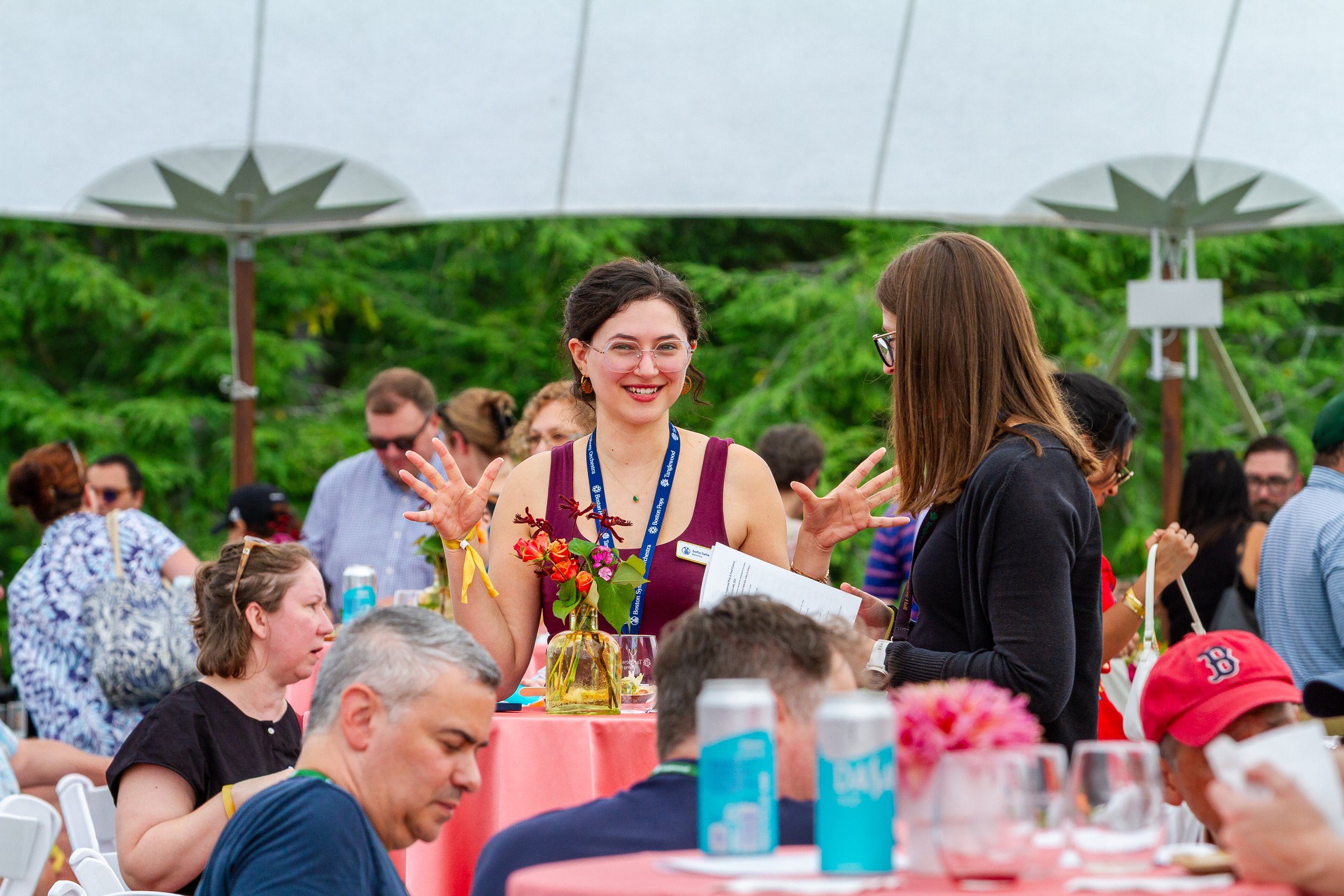 A woman with glasses and earrings smiling and gesturing with her hands, standing outdoors among a group of people at a social gathering or event, with green trees and a large shade in the background.