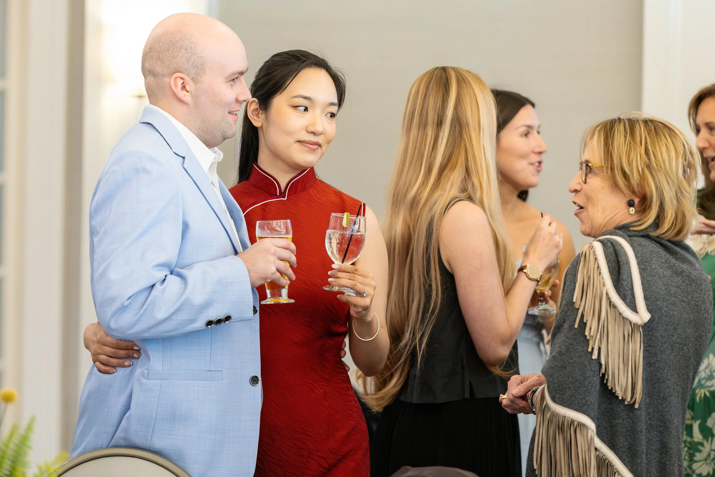 Group of people at a social event, holding drinks and engaging in conversation, dressed in formal and semi-formal attire.
