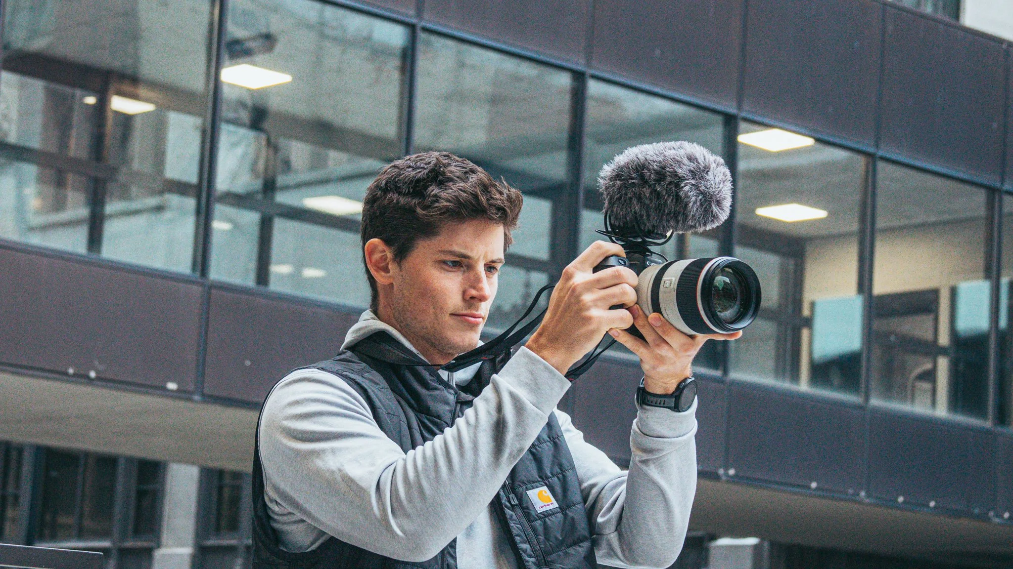 A young man is filming with a professional video camera outdoors in front of a modern glass building.
