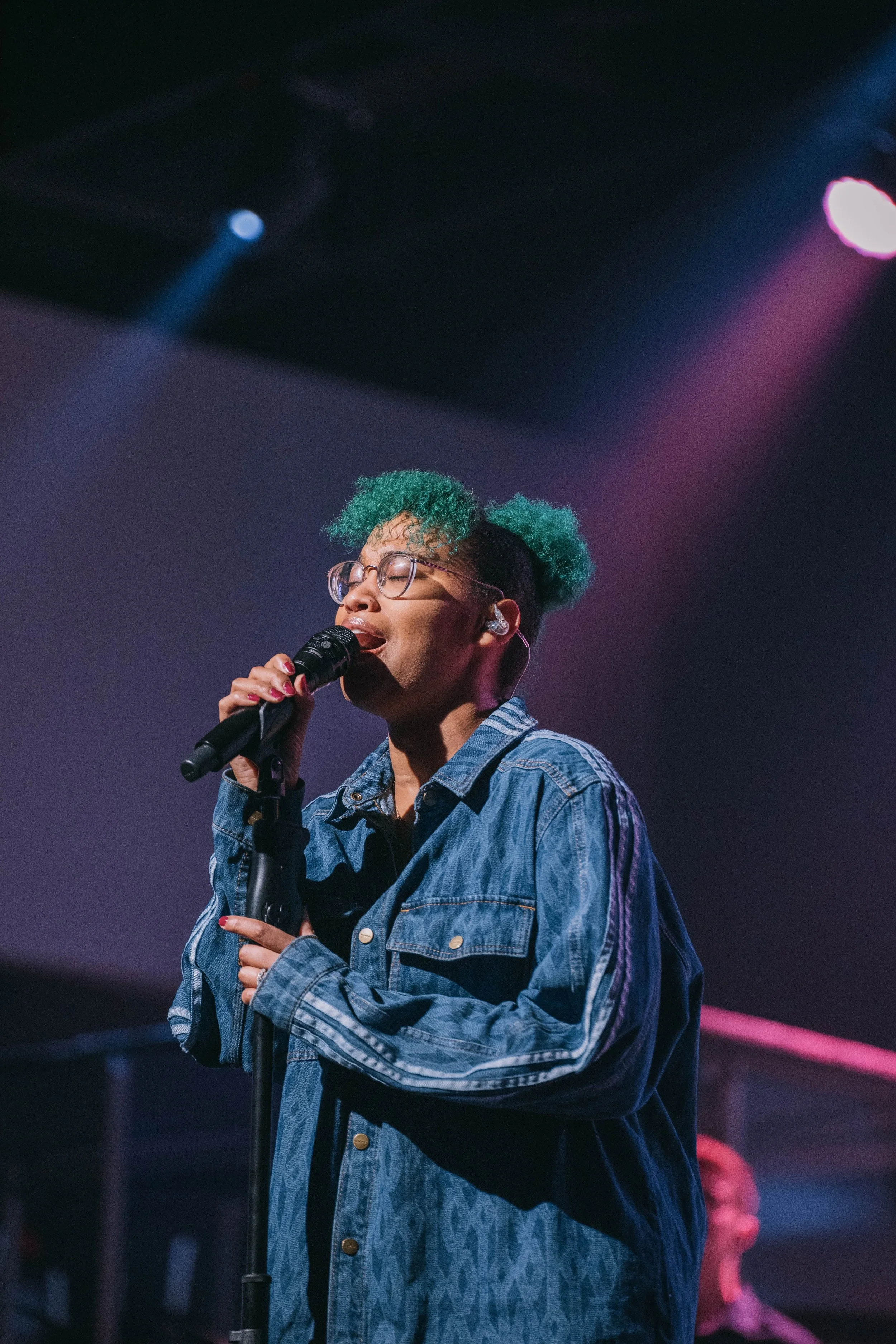 A person singing into a microphone on stage, wearing glasses and a denim jacket, with green hair styled in two puffs and a blurred background with stage lights.