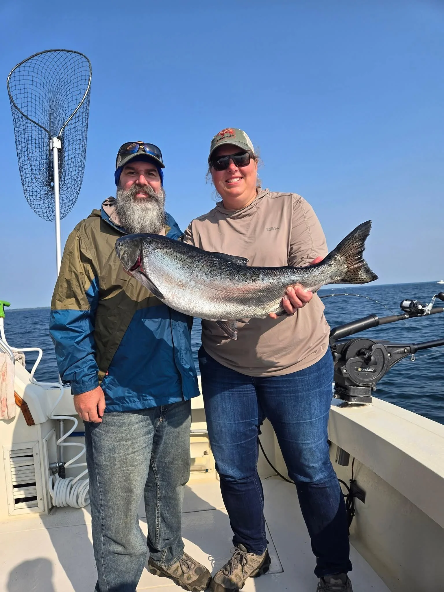 A man and woman on a boat holding a large fish they caught, with a fishing net and ocean in the background during daytime.