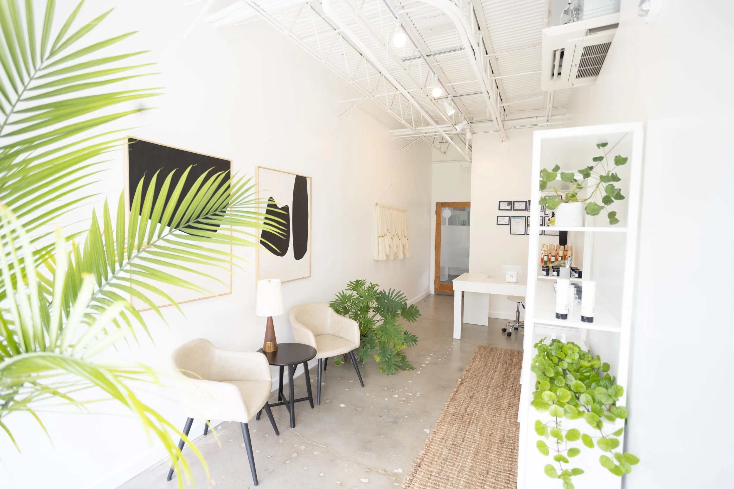 Bright minimalist interior with white walls, modern furniture, and green plants including a large palm leaf, with framed black and white artwork on the wall, a small desk area, and a wooden doorway.