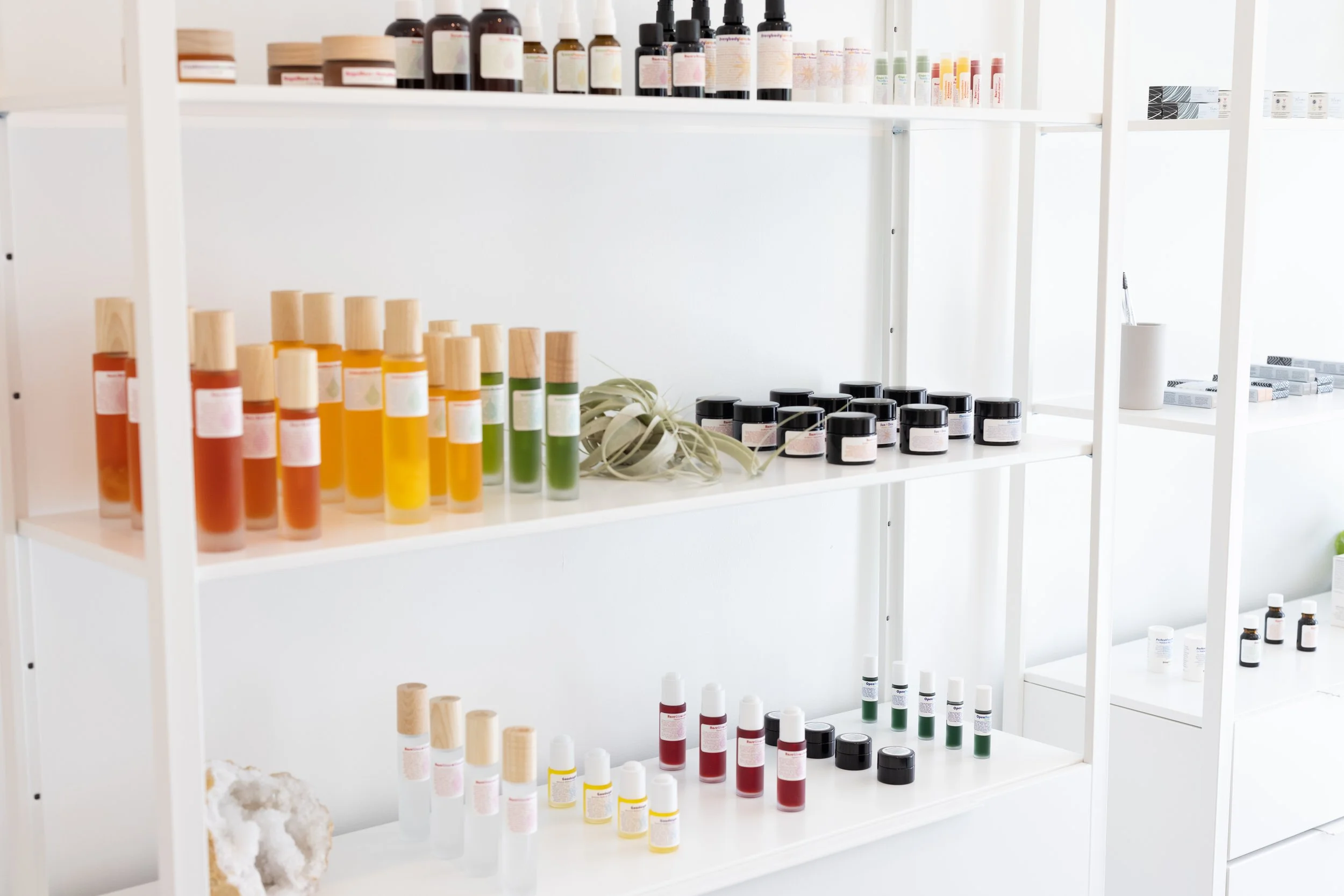 White shelves filled with various cosmetic products, including bottles and jars with black, white, and wooden caps, and small tubes, organized in rows.