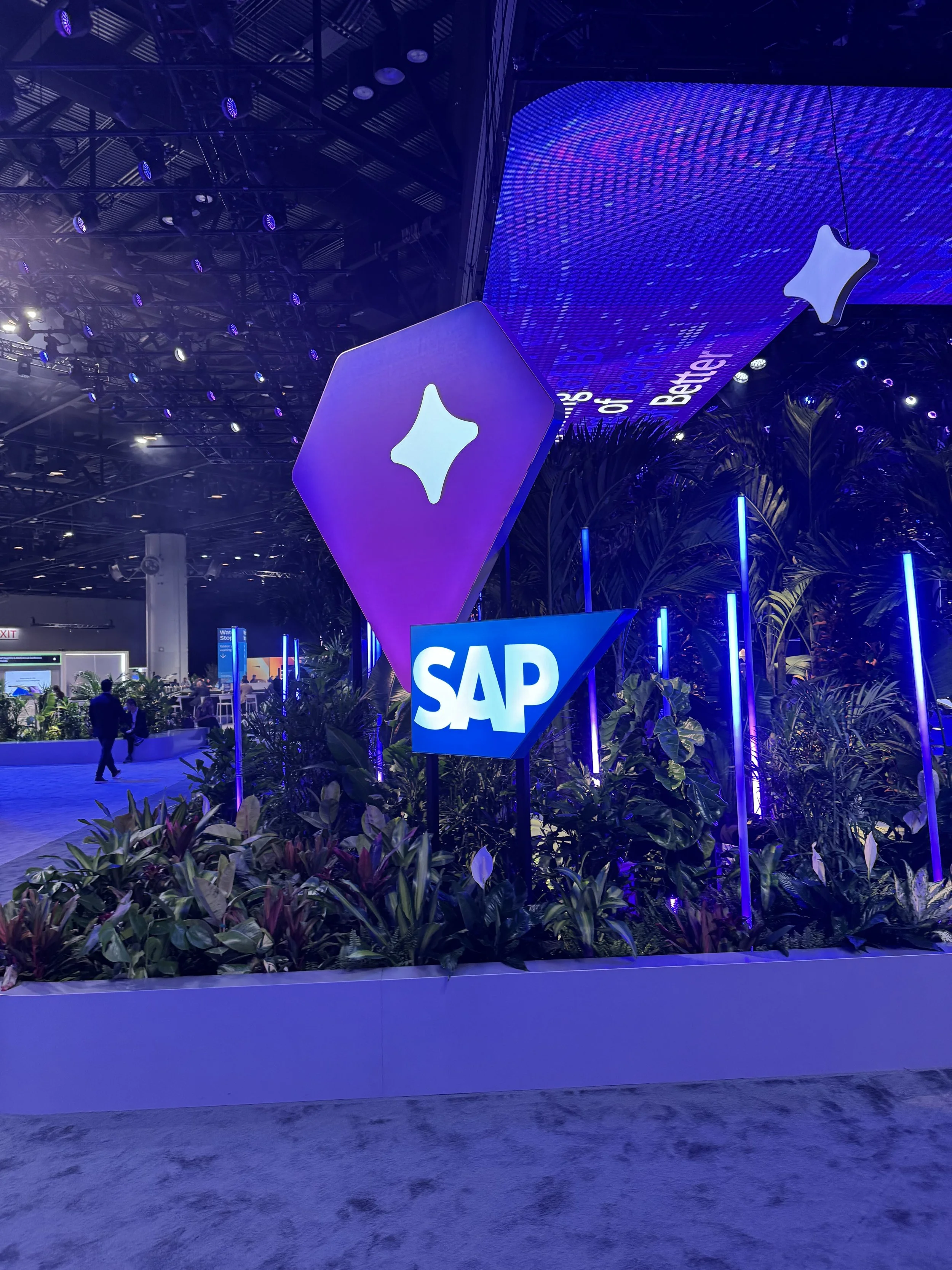 Indoor event space with blue and purple lighting, displaying SAP and Apple logos, decorated with lush green plants and modern lighting fixtures.